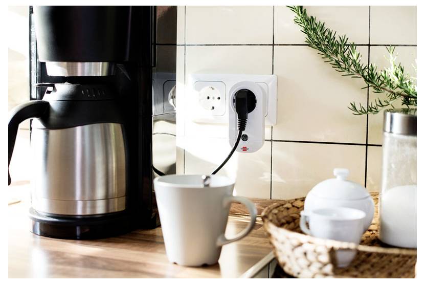 Kaffeemaschine und Becher, die in eine Wandsteckdose auf einer Küchenarbeitsplatte eingestöpselt sind, mit Bohnen und Geschirr in der Nähe.