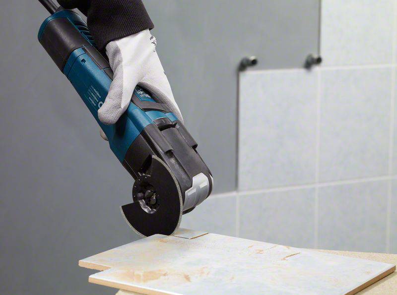 Hand mit Handschuh hält ein elektrisches Schneidewerkzeug, das eine Keramikfliese schneidet. Hintergrund zeigt eine geflieste Wand.