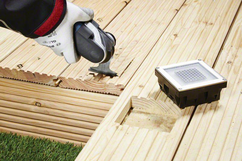 Eine Person installiert mit einem Werkzeug eine Holzterrasse und eine quadratische Einbauleuchte.