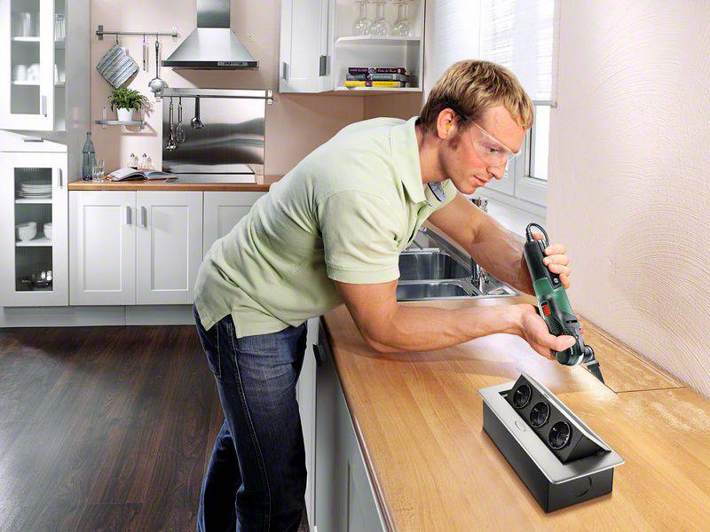 Ein Mann arbeitet mit einem Elektrowerkzeug auf einer Küchenarbeitsplatte. Er trägt eine Schutzbrille und steht in einer modernen Küche.