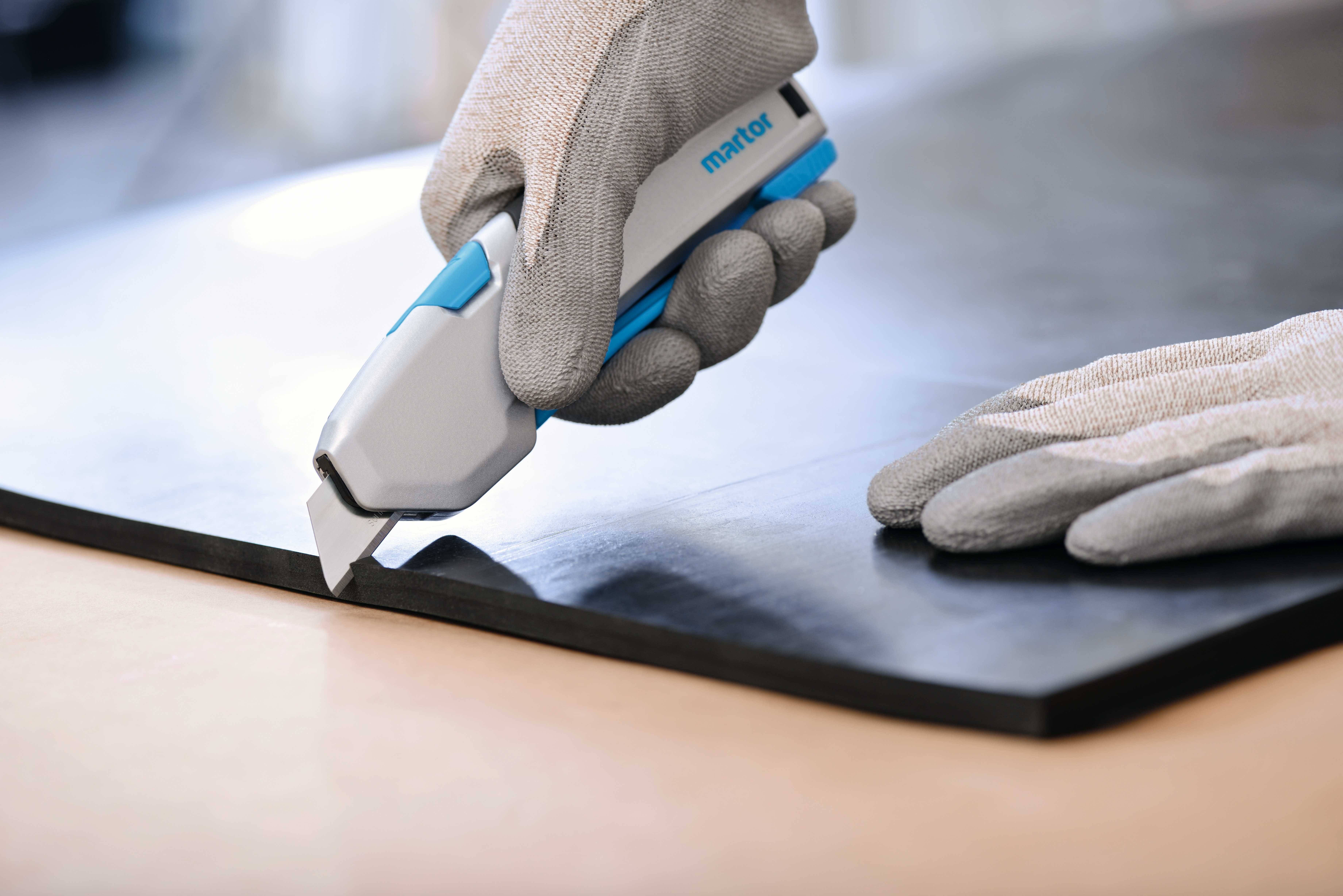 Eine Person schneidet mit einem Sicherheitsmesser eine schwarze Matte. Beide Hände tragen graue Arbeitshandschuhe.