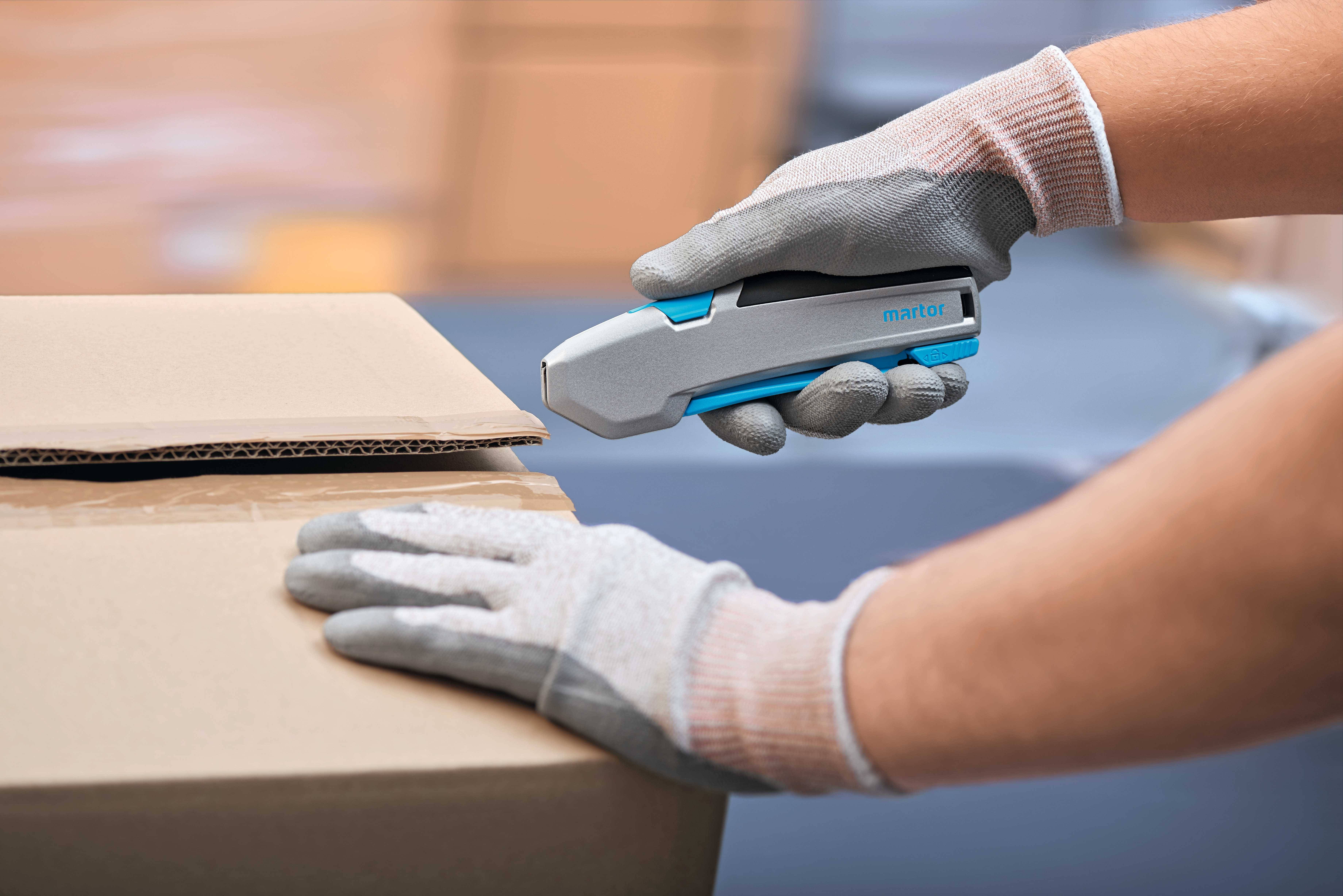 Person mit Handschuhen öffnet vorsichtig einen Karton mit einem grauen Schneidewerkzeug. Hintergrund unscharf.