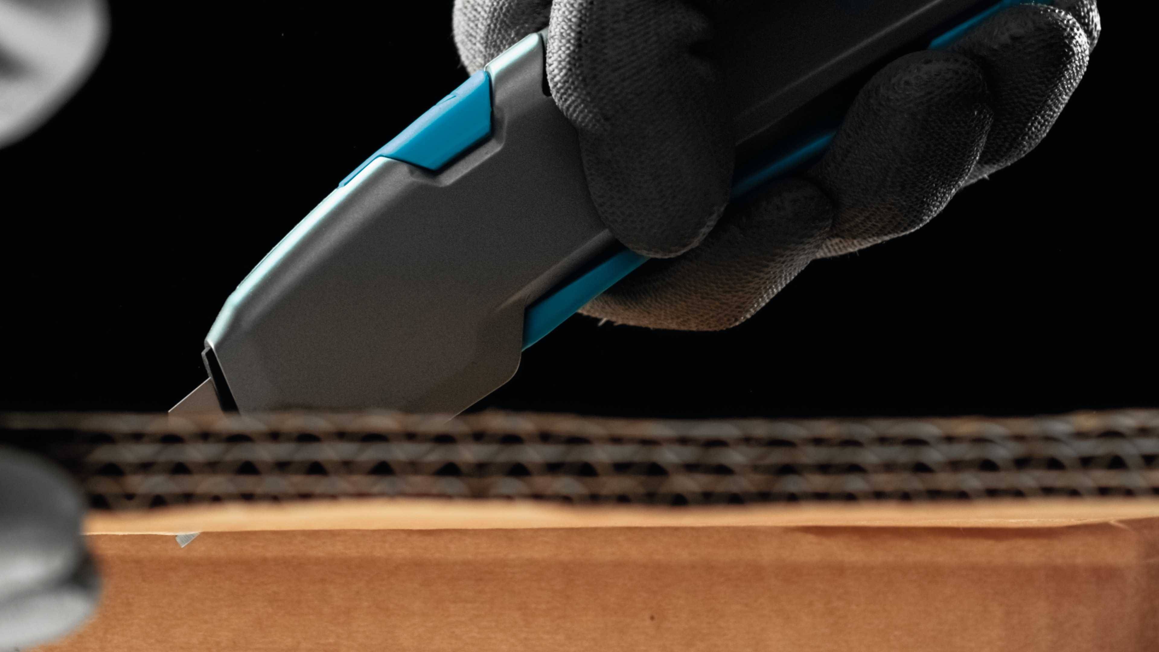 Eine Hand in einem Handschuh schneidet einen Karton mithilfe eines grauen und blauen Cuttermessers.