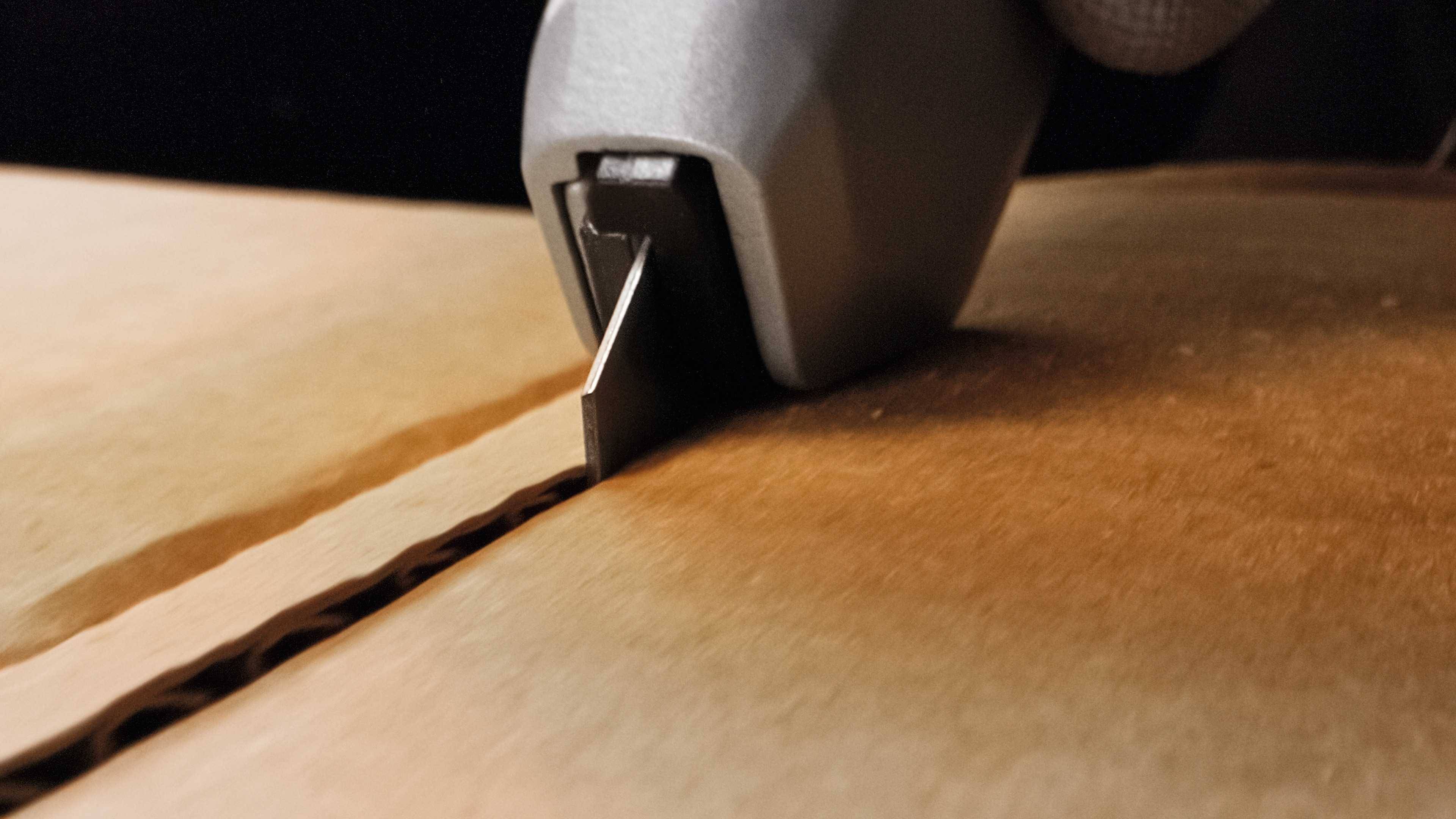 Nahaufnahme einer Hand, die mit einem Cuttermesser einen Karton öffnet. Der Fokus liegt auf der Klinge und der Schnittlinie im Karton.