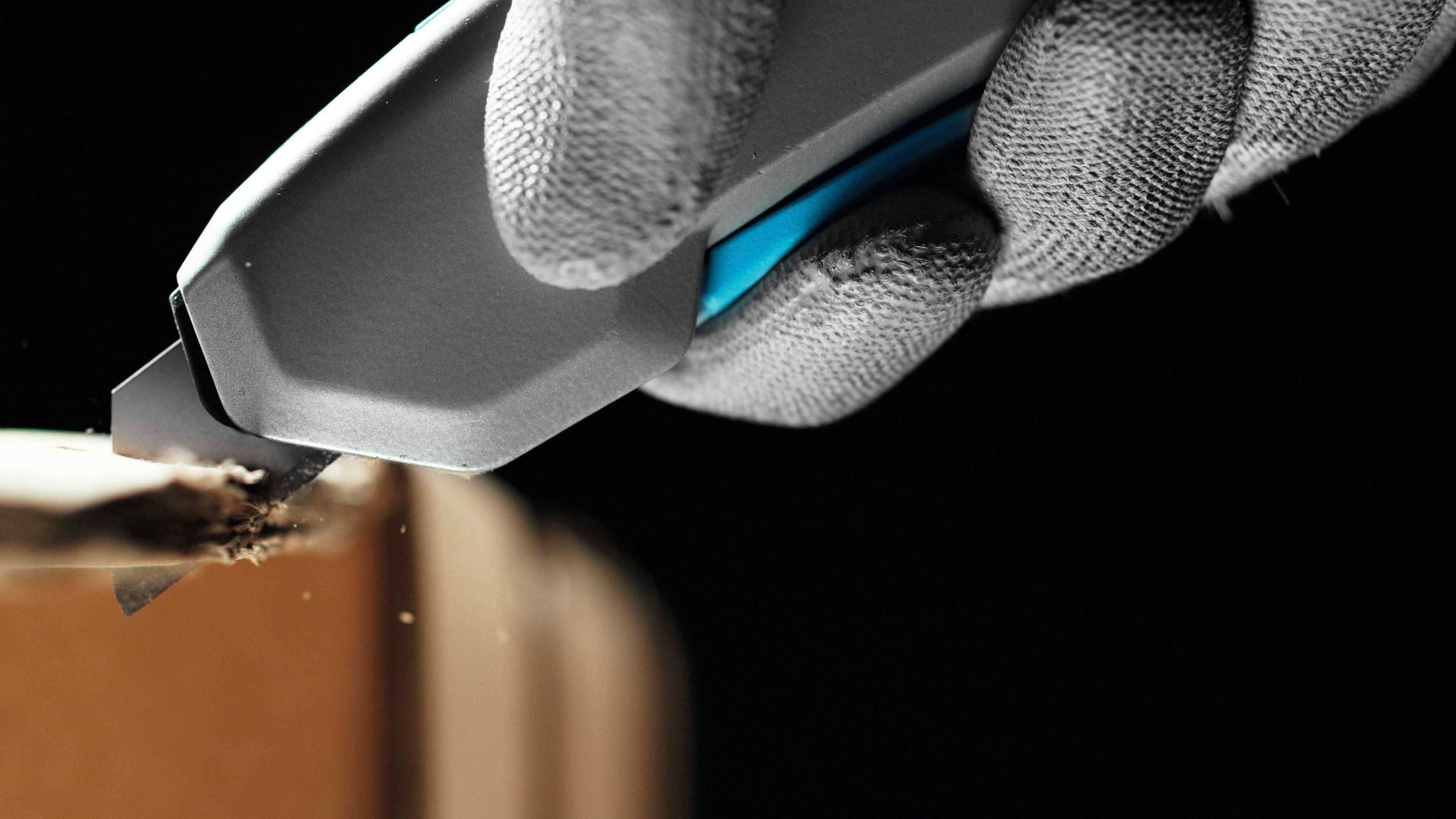 Eine behandschuhte Hand schneidet mit einem grauen und blauen Schneidemesser durch Pappe.