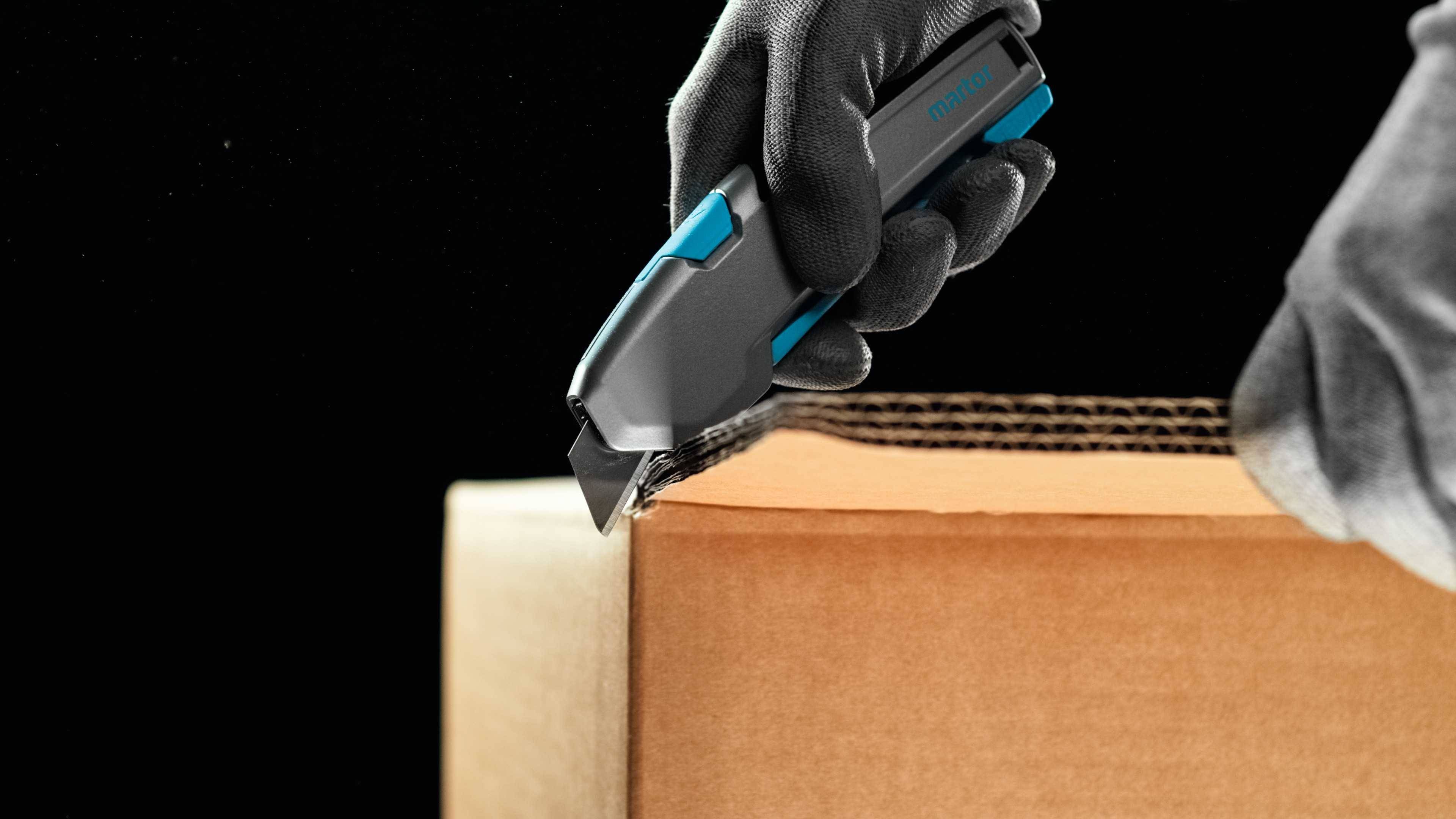 Eine bekleidete Hand schneidet mit einer Teppichmesserklinge einen Pappkarton auf.
