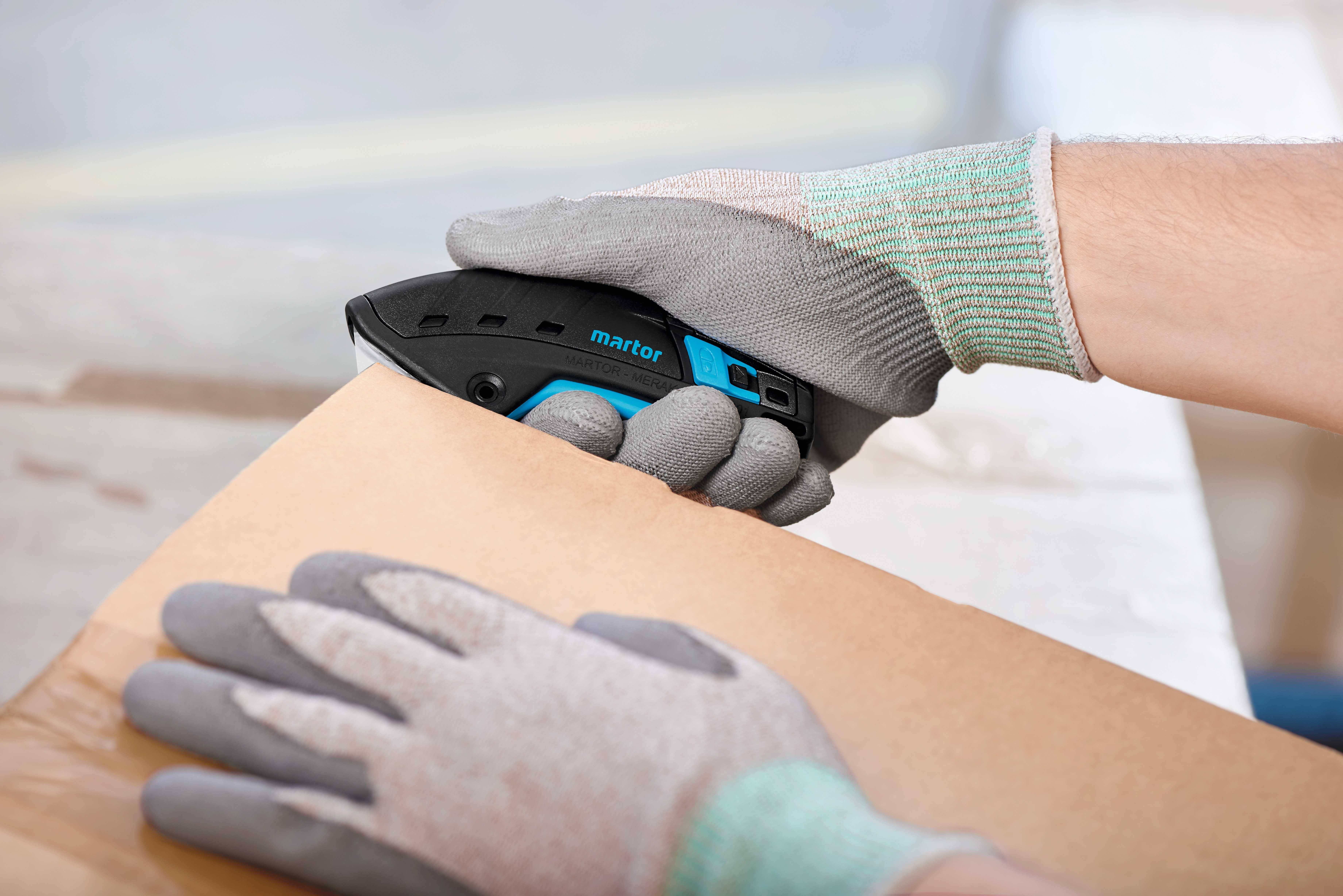 Person mit Schutzhandschuhen schneidet ein Paket mit einem Sicherheitsmesser auf einer Werkbank.