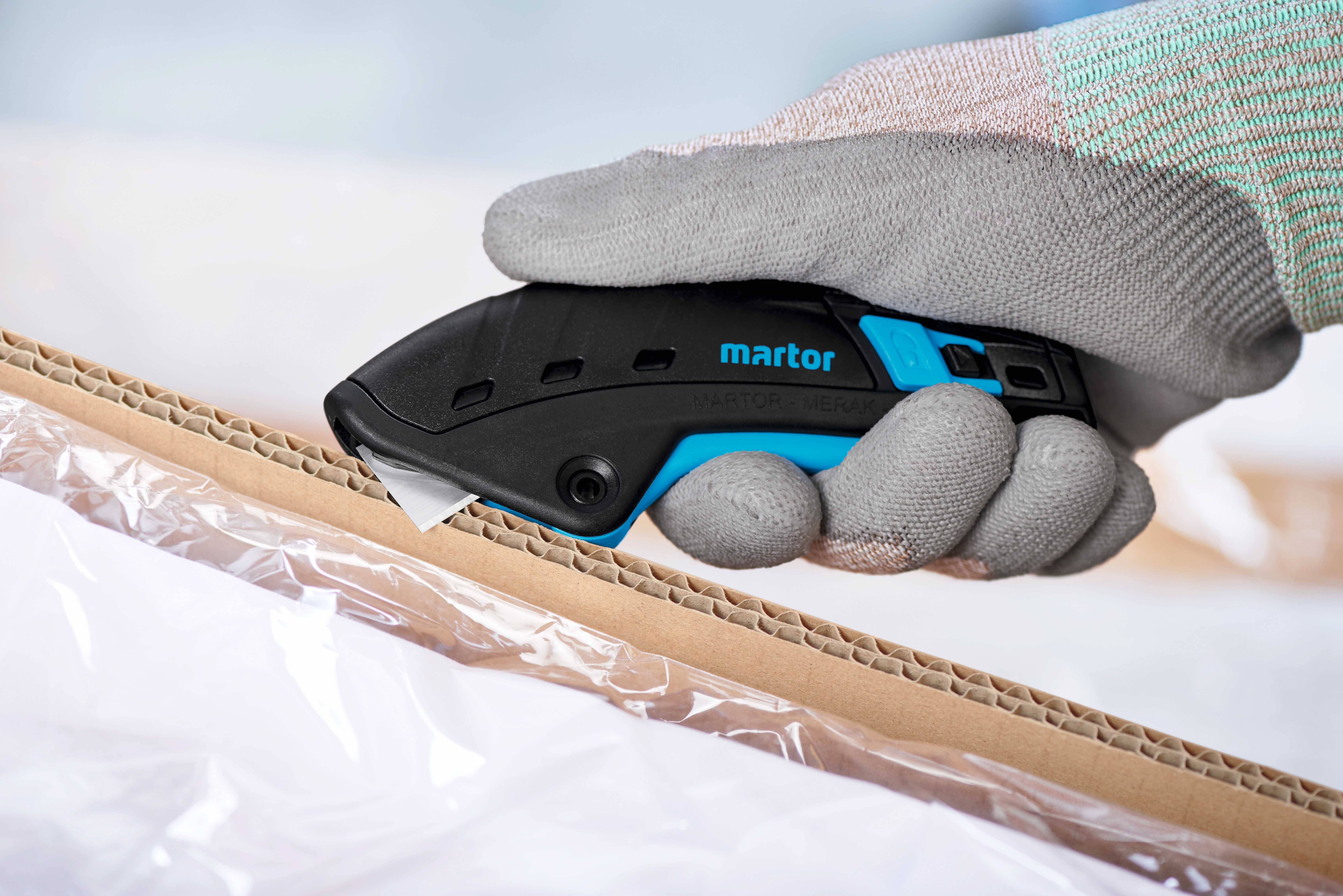 Mitarbeiter mit grauen Handschuhen verwendet ein Martor-Sicherheitsmesser, um einen Karton sorgfältig zu öffnen; transparentes Plastik umhüllt.