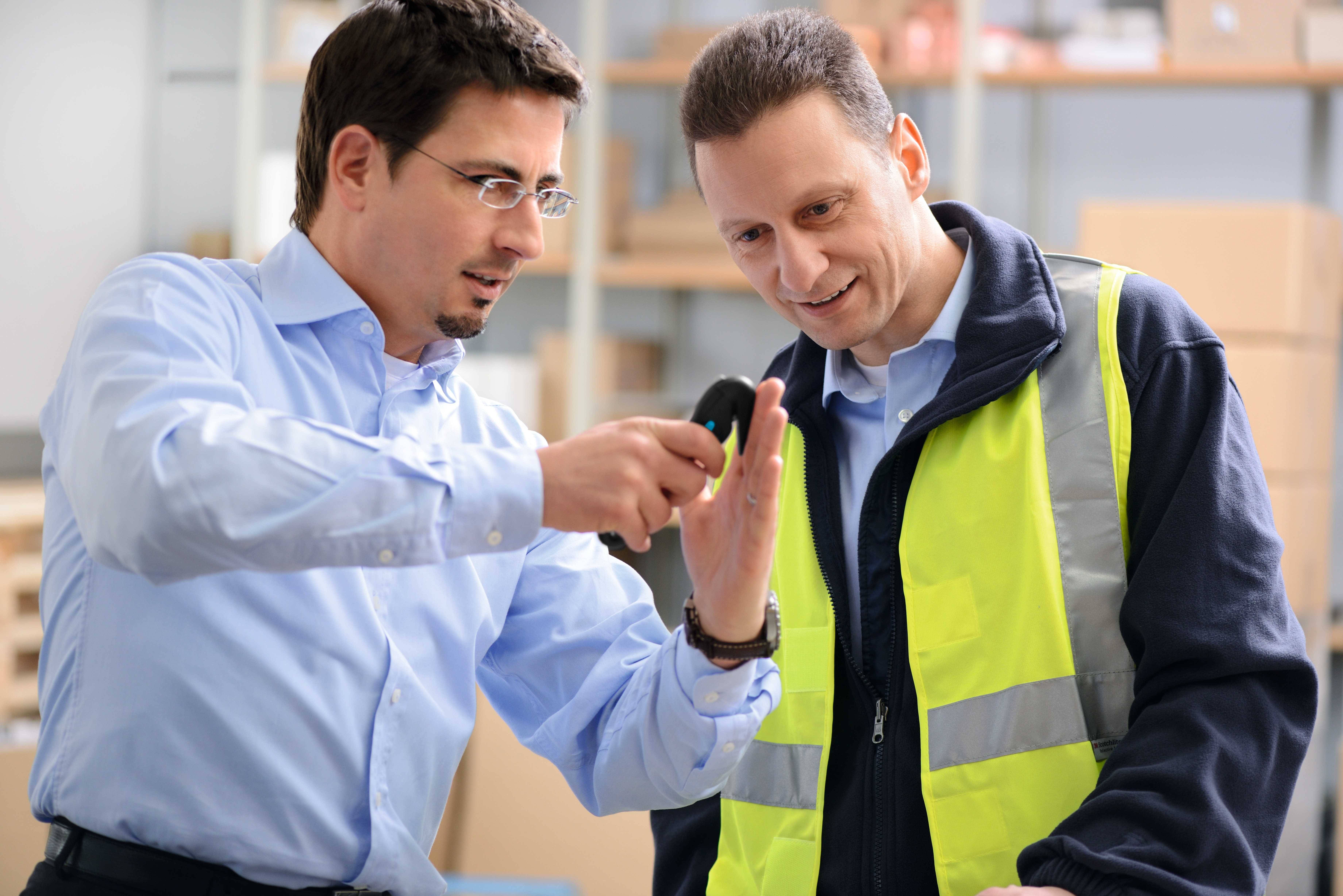 Zwei Männer in einem Lagerraum; der eine im Hemd zeigt dem anderen im Warnschutz eine kleine elektronische Vorrichtung.