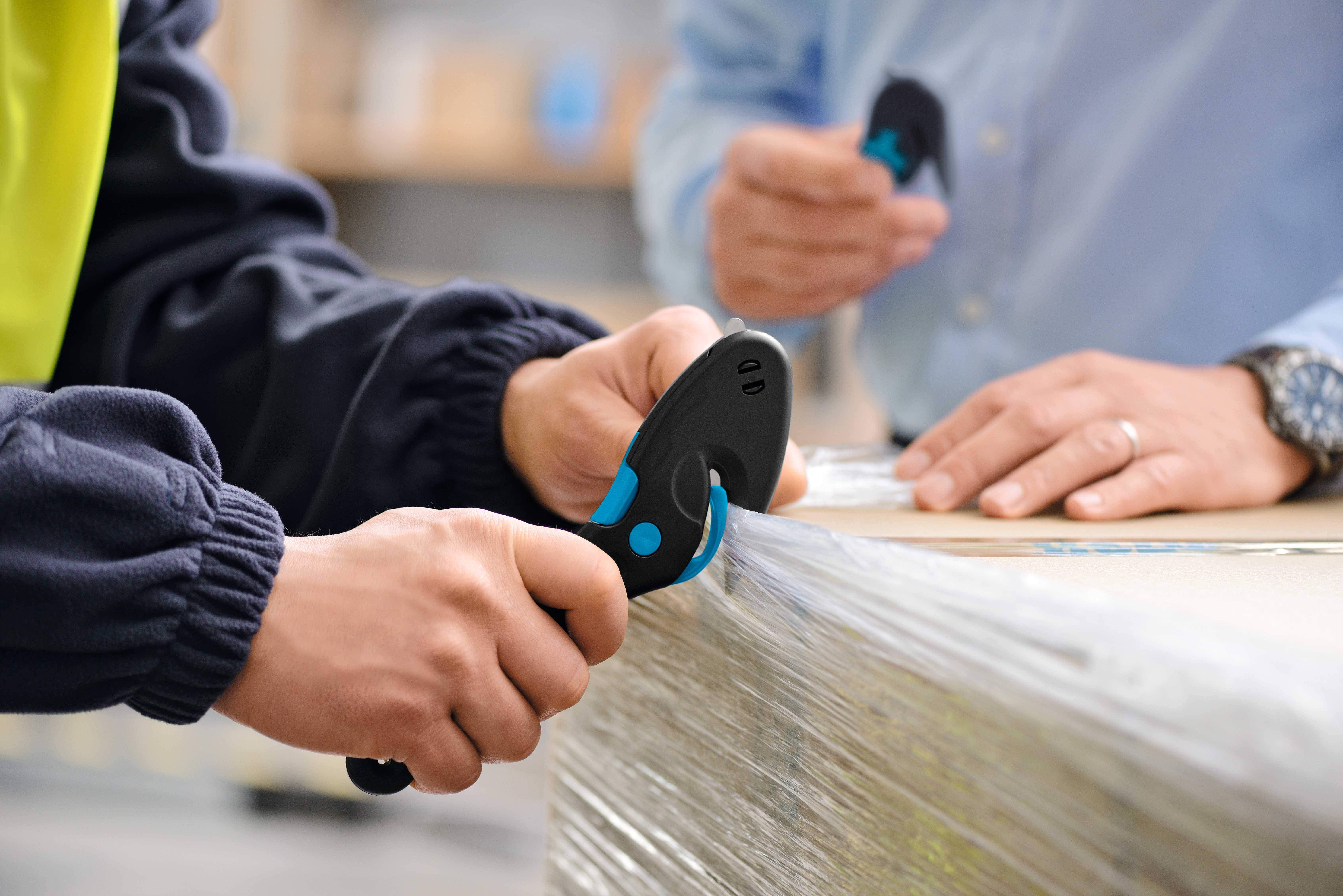 Zwei Personen öffnen eine Kartonverpackung mit Sicherheitsschneidern; eine trägt eine gelbe Arbeitsweste und die andere ein blaues Hemd.