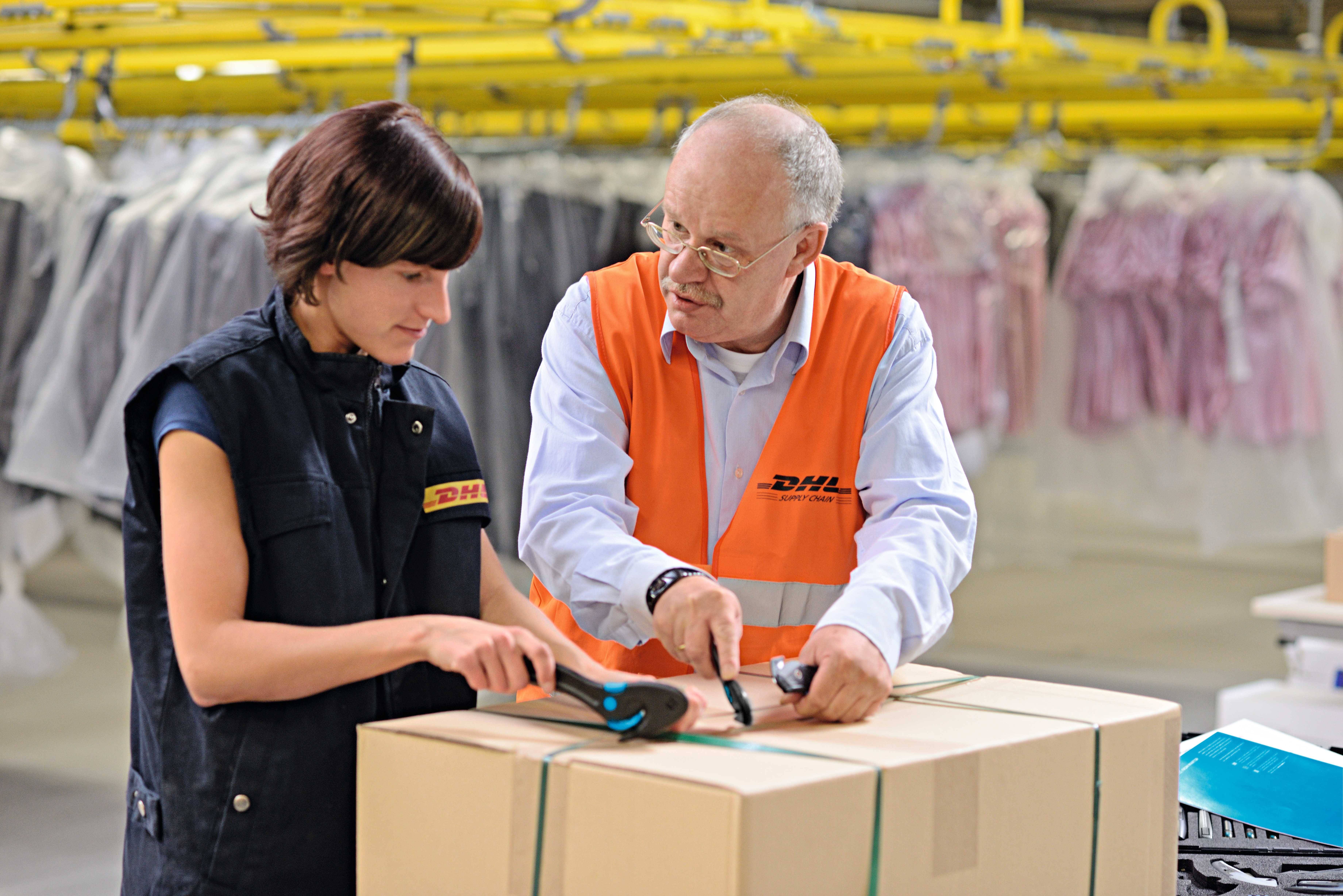 Zwei Personen in DHL-Uniform arbeiten gemeinsam in einem Lager an einem Paket. Im Hintergrund sind Kleidungsstücke sichtbar.