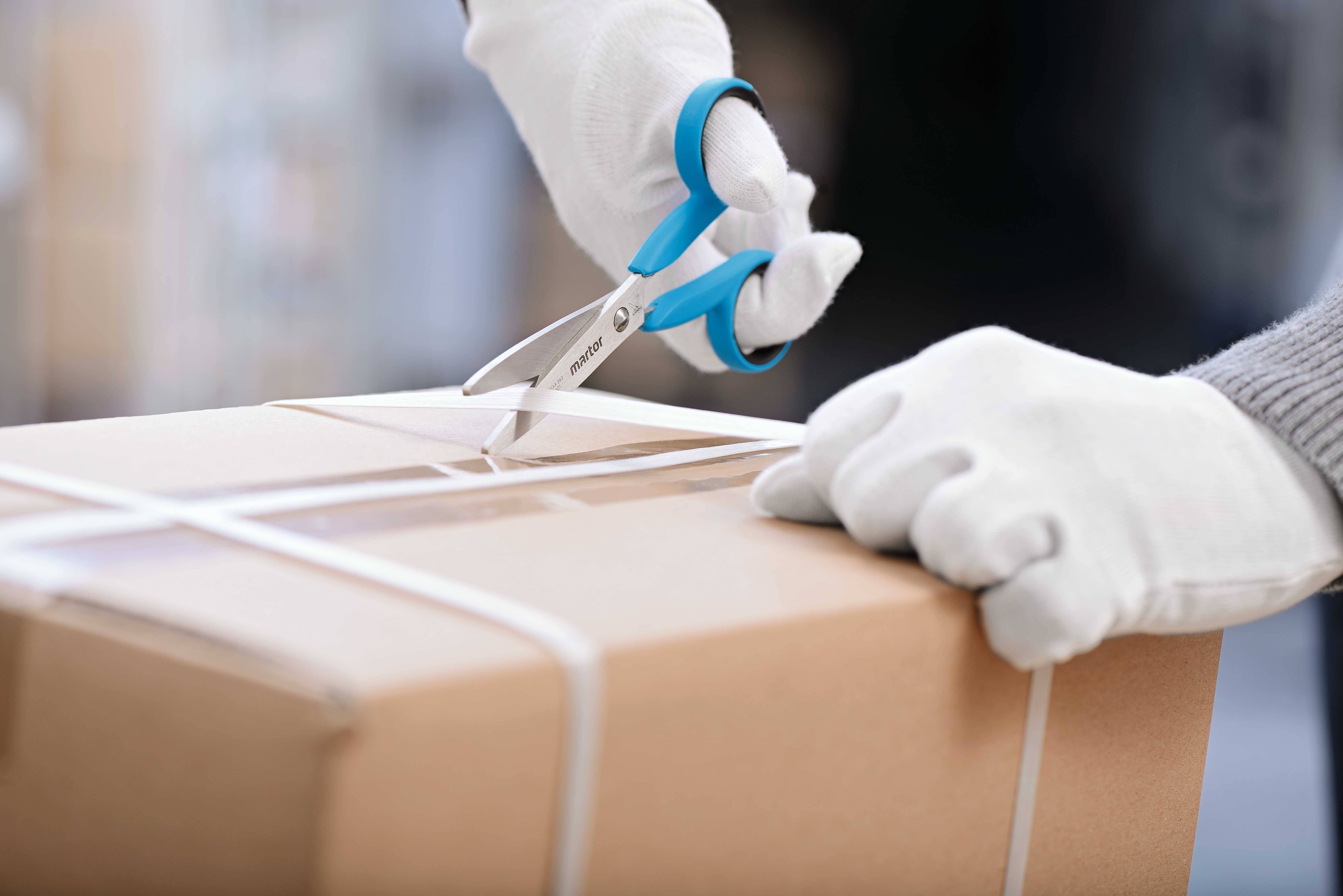 Eine Person schneidet mit einer Schere ein Paketband von einem Karton. Die Hände tragen weiße Handschuhe.