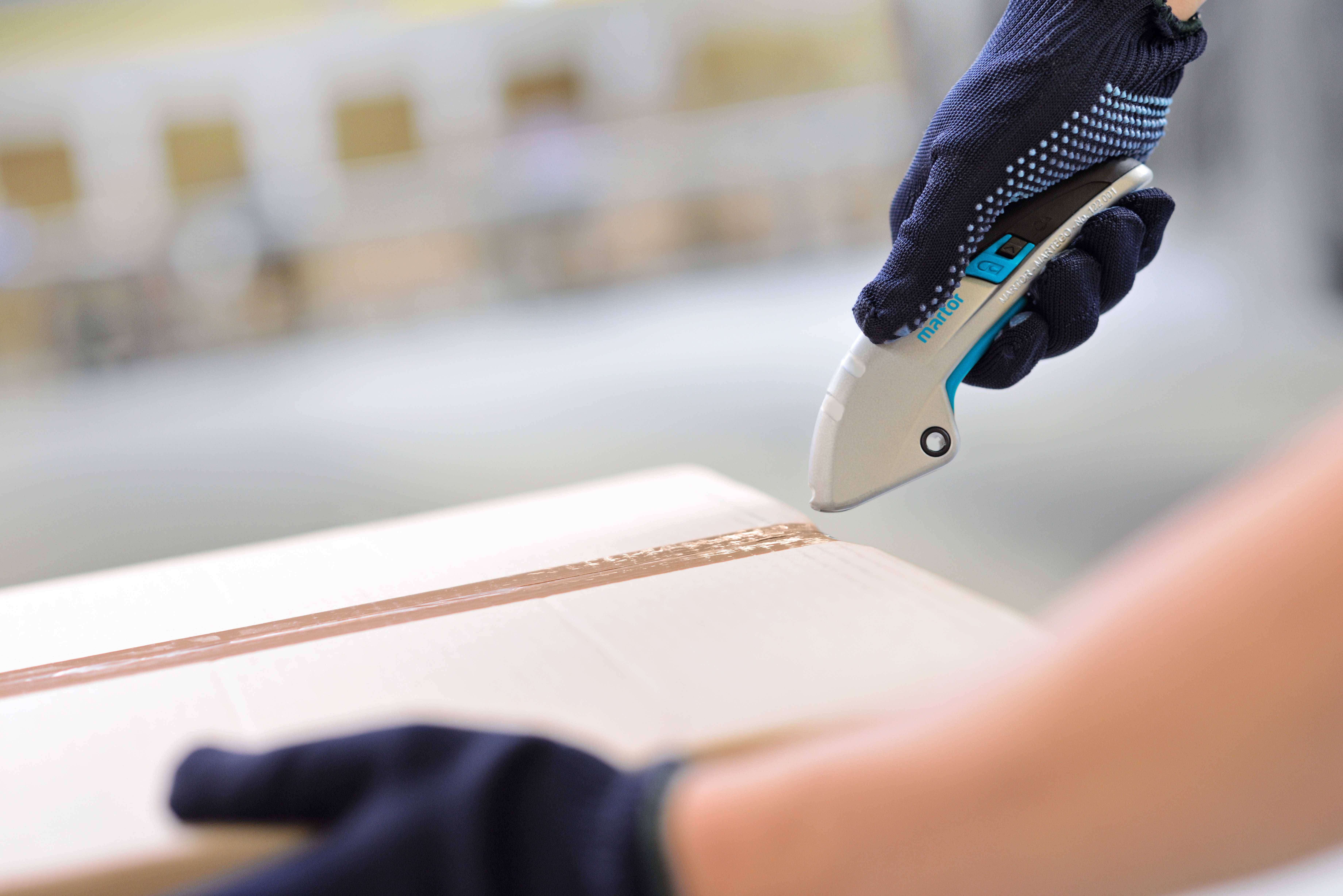 Eine Person öffnet mit einem Cuttermesser ein Paket. Sie trägt blaue Handschuhe und schneidet das Klebeband auf.
