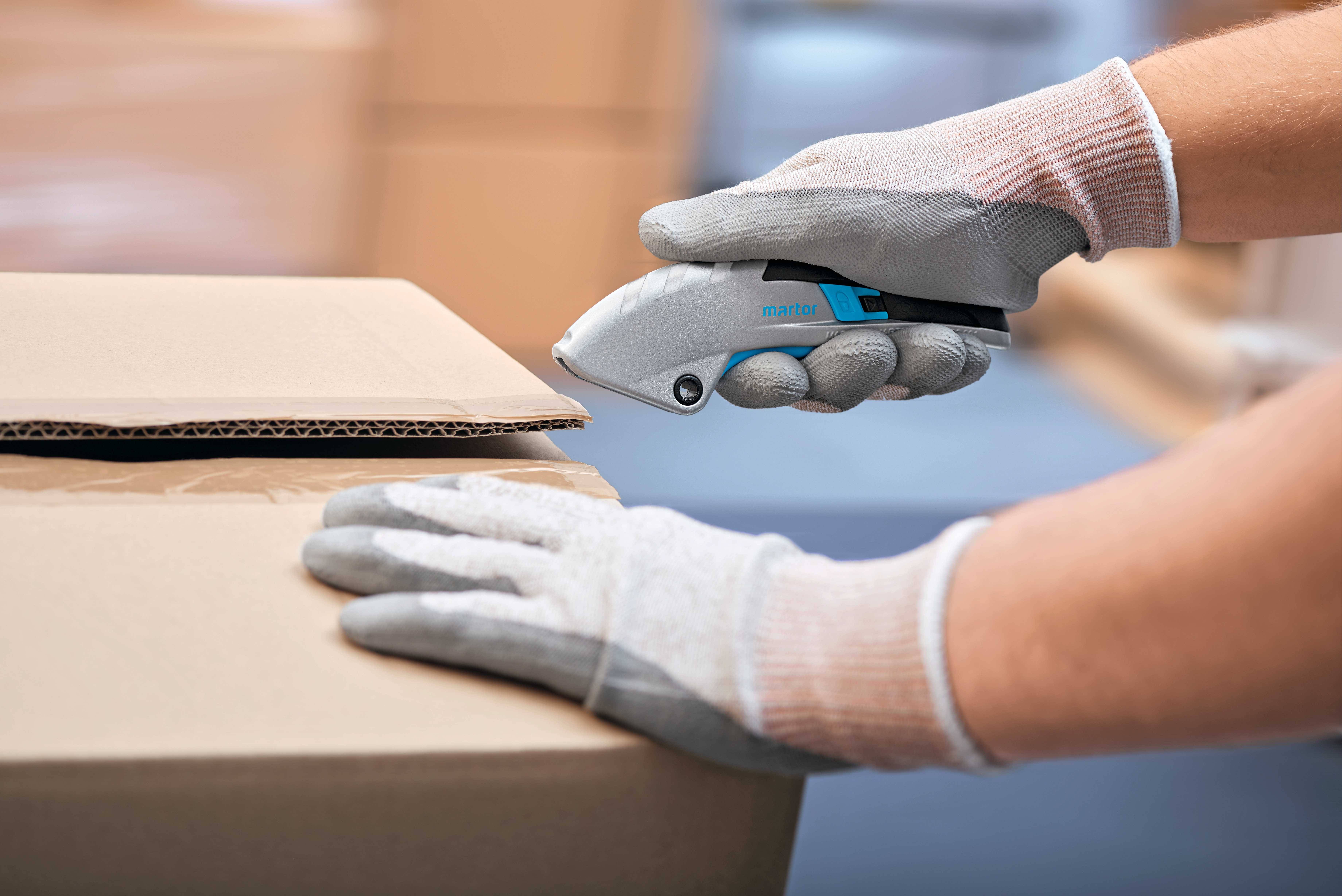 Eine Person schneidet mit einem Cuttermesser einen Karton. Sie trägt graue Handschuhe und öffnet vorsichtig den oberen Deckel.