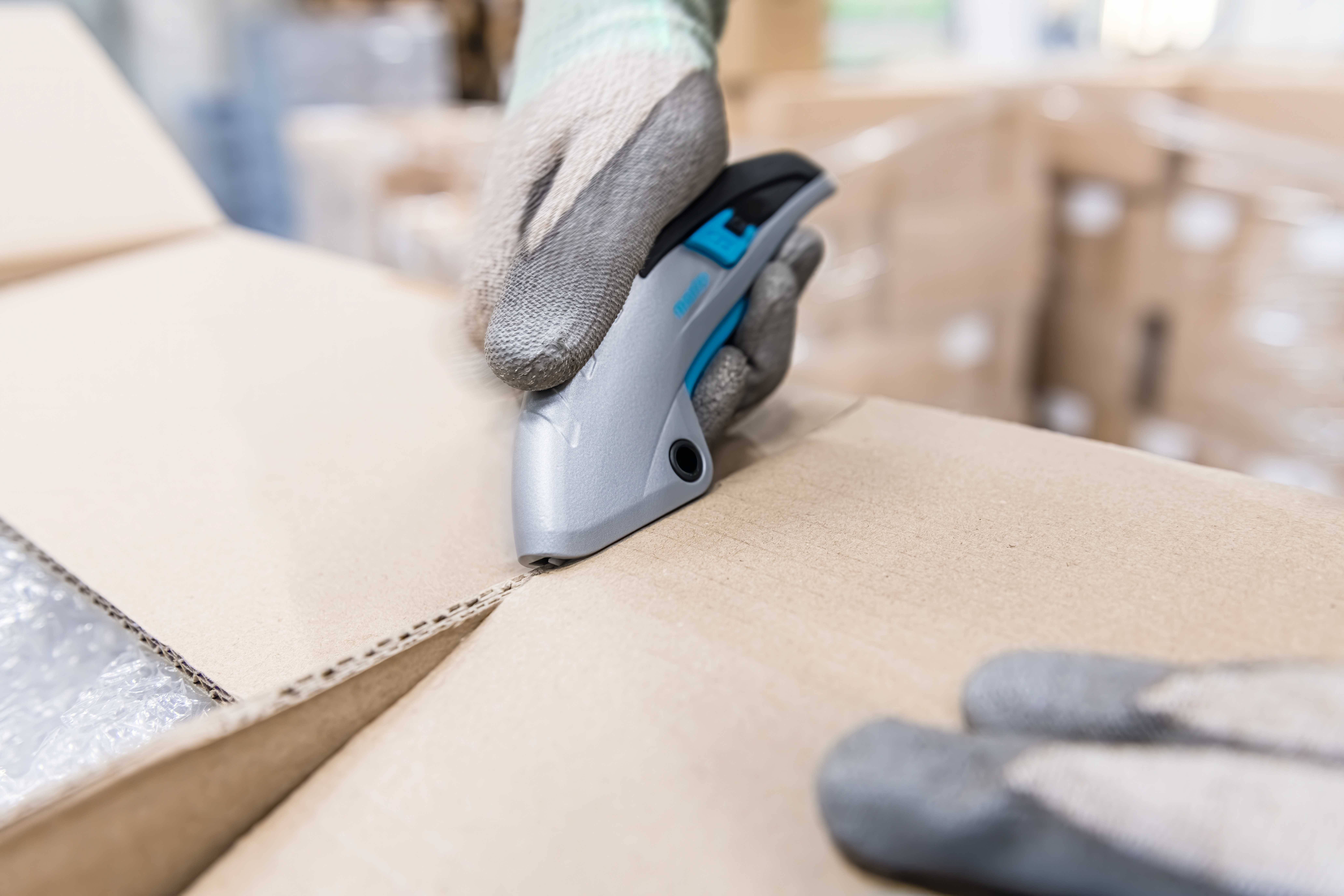 Eine Person schneidet mit einem Sicherheitsmesser ein Kartonpaket auf. Sie trägt Handschuhe. Im Hintergrund sind weitere Pakete zu sehen.