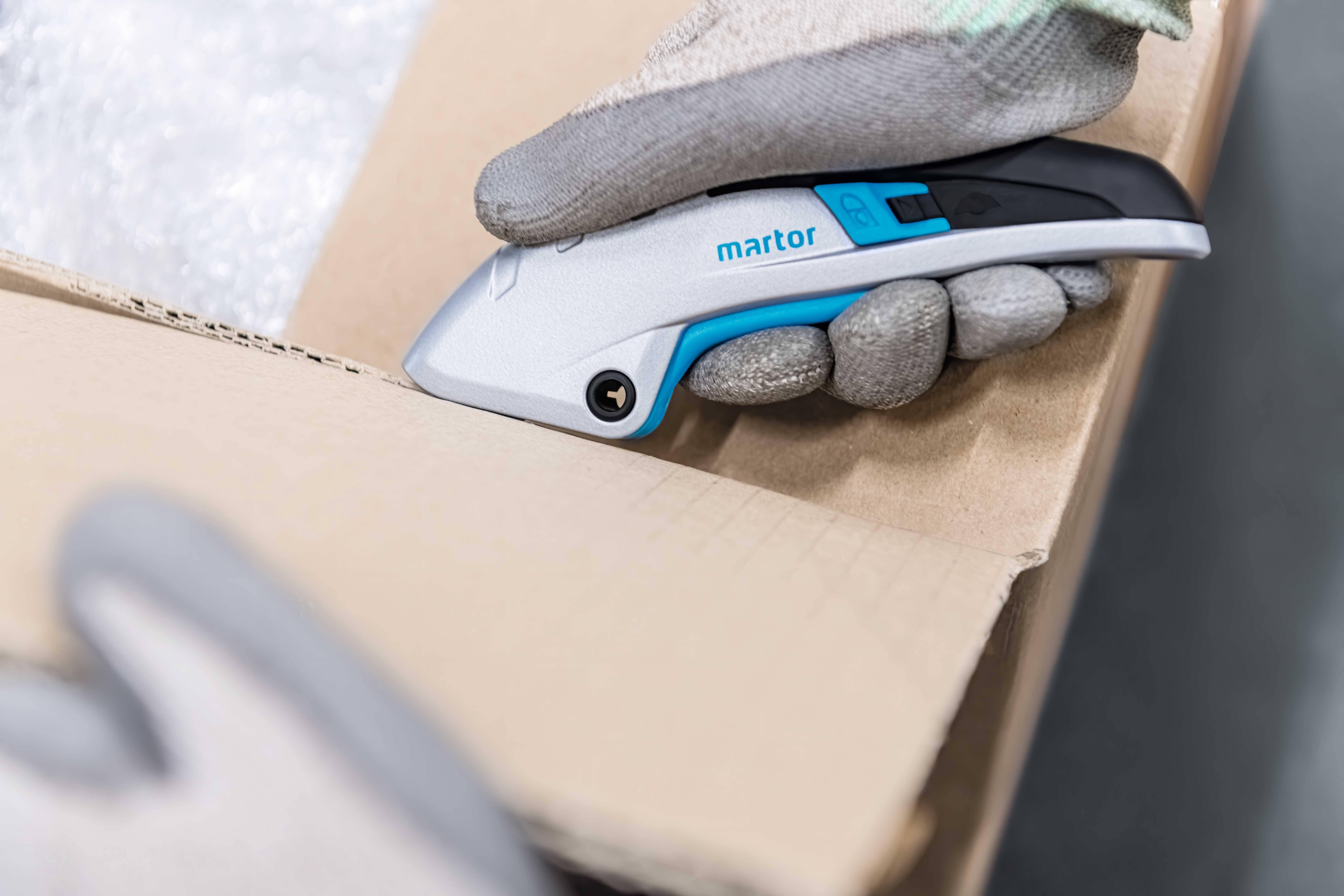 Eine Hand mit Handschuh nutzt einen Sicherheitsschneider, um einen Karton zu öffnen. Der Schneider ist silbern und blau.