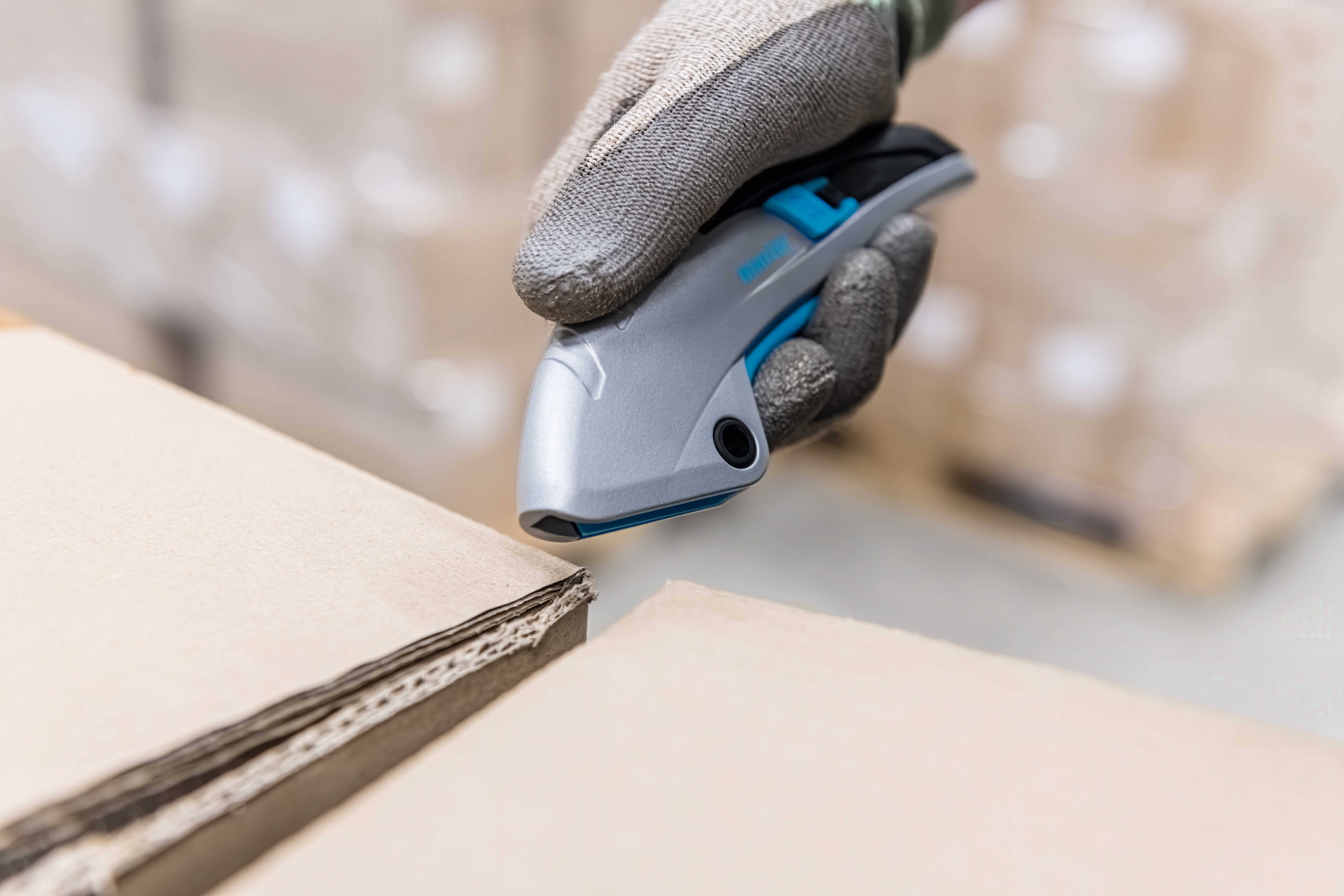 Eine behandschuhte Hand verwendet ein Sicherheitsmesser zum Schneiden eines Kartons. Umgebung unscharf, Fokus auf Messer und Karton.