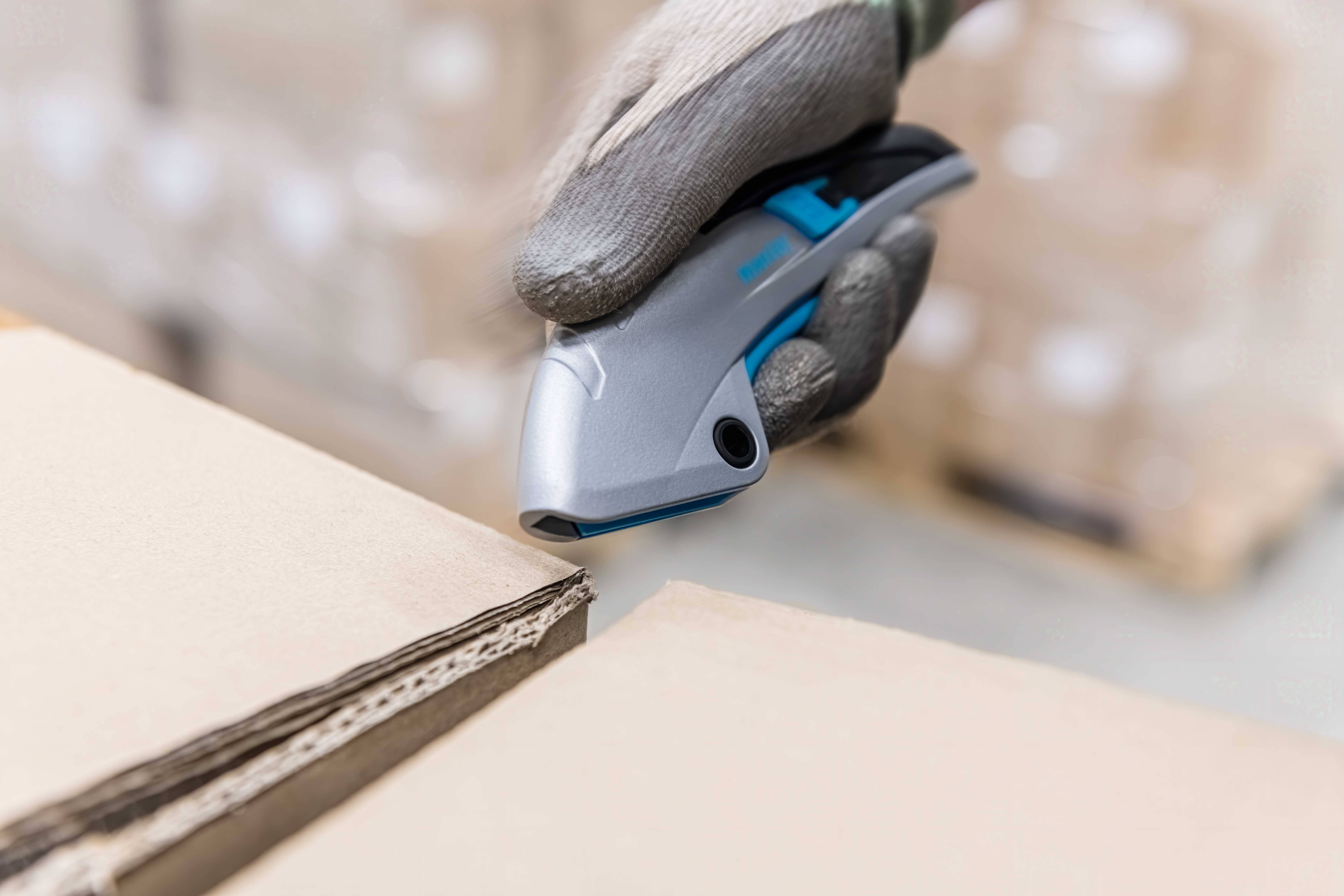 Hand in Handschuhen nutzt ein Verpackungsmesser, um einen Karton zu öffnen. Kartons im Hintergrund deuten auf Lagerumgebung hin.
