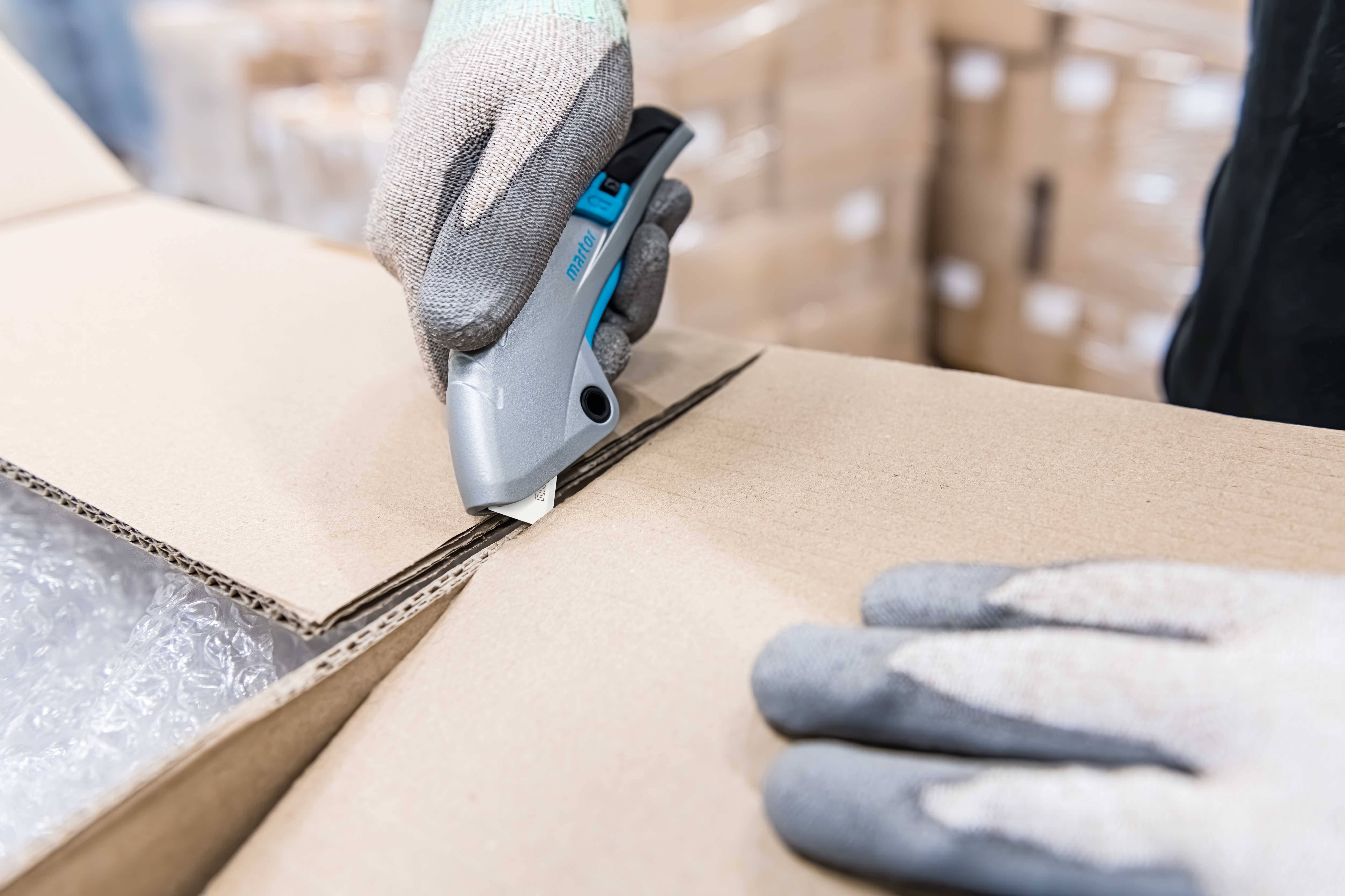 Eine Person schneidet mit einem grauen Cuttermesser einen Karton. Der Karton enthält Plastikfolienverpackungen. Hände tragen Arbeitshandschuhe.