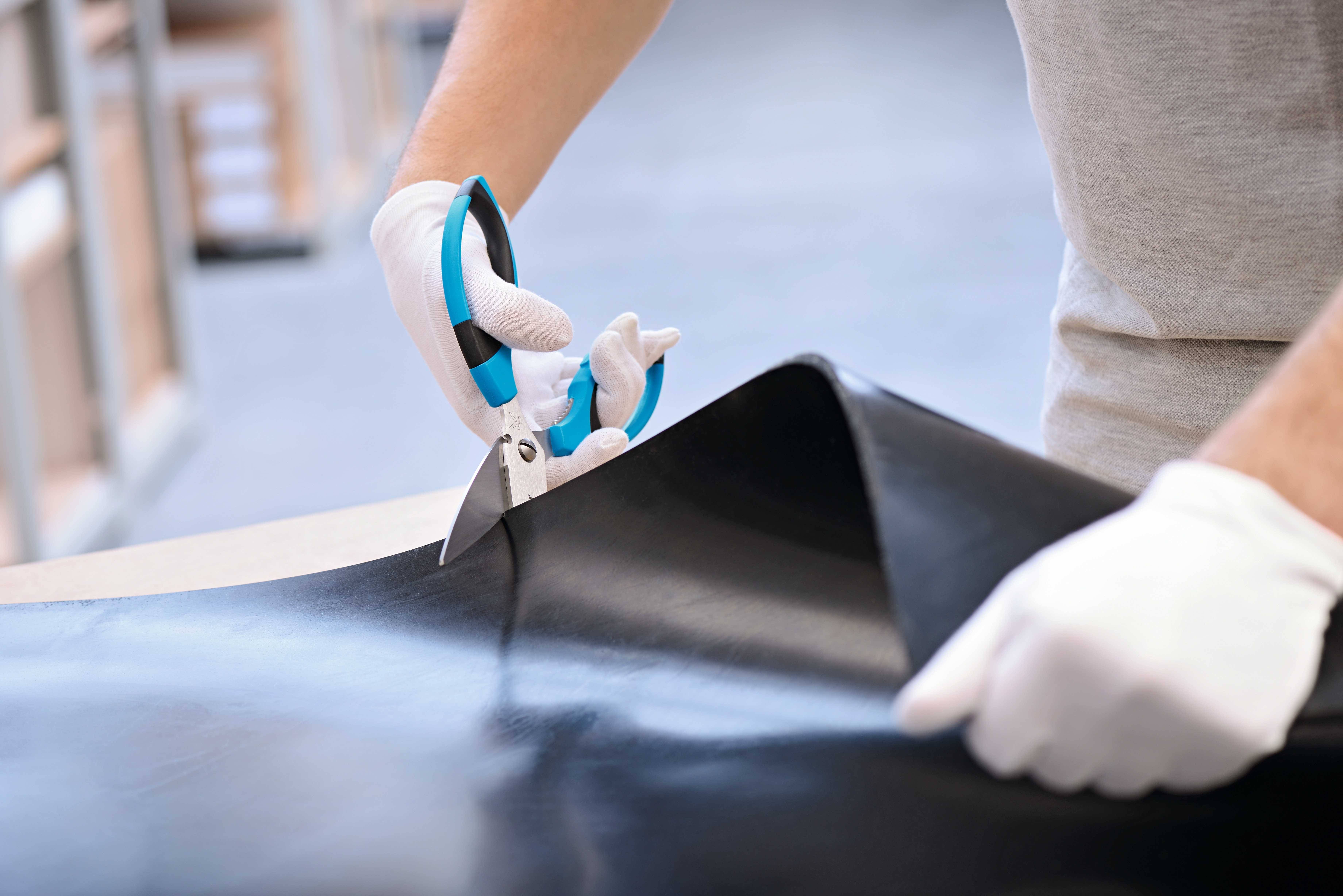 Eine Person mit weißen Handschuhen schneidet mit einer blauen Schere ein großes schwarzes Lederstück auf einem Tisch.