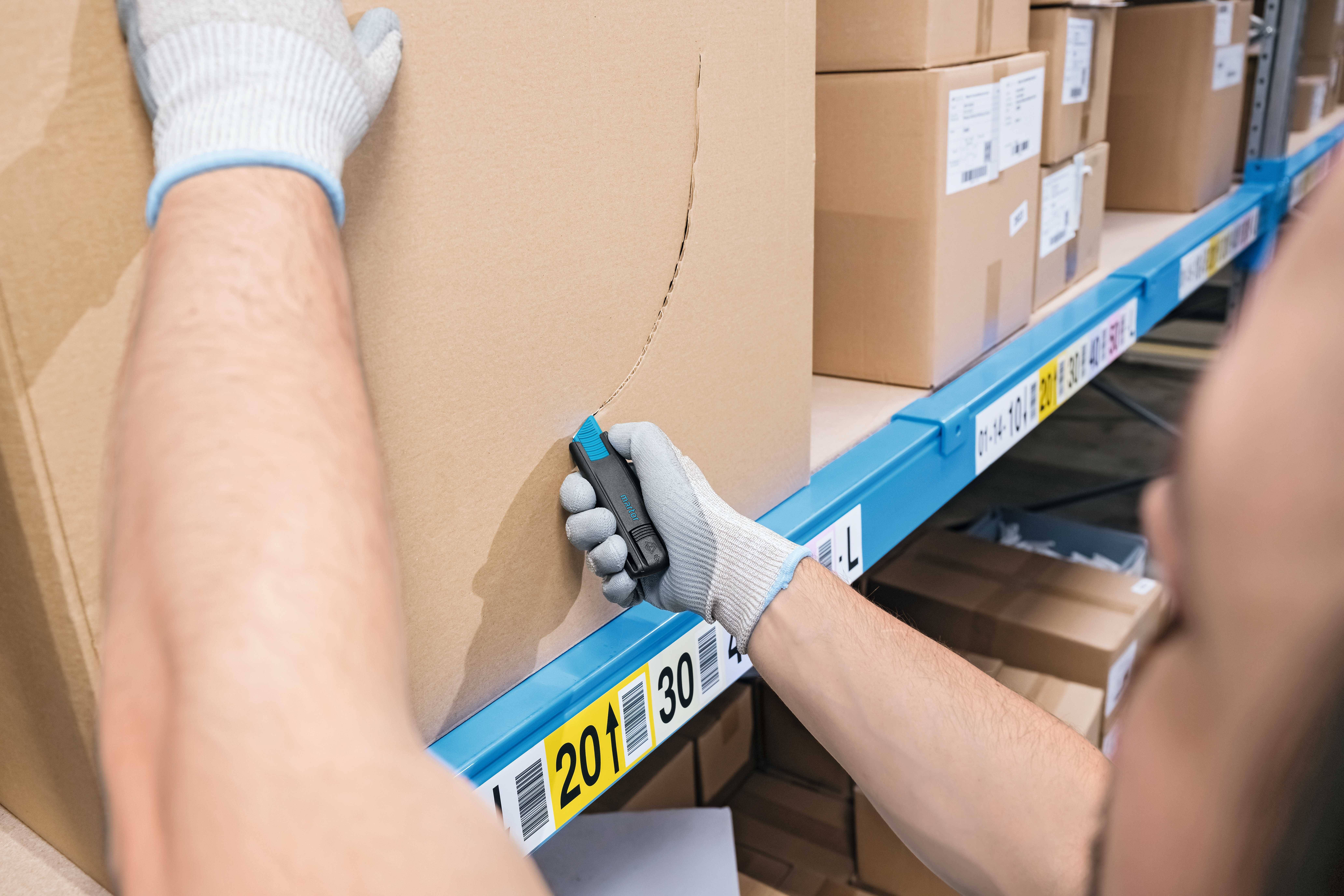 Eine Person öffnet einen großen Pappkarton mit einem Cutter-Messer in einem Lager, während sie Schutzhandschuhe trägt.