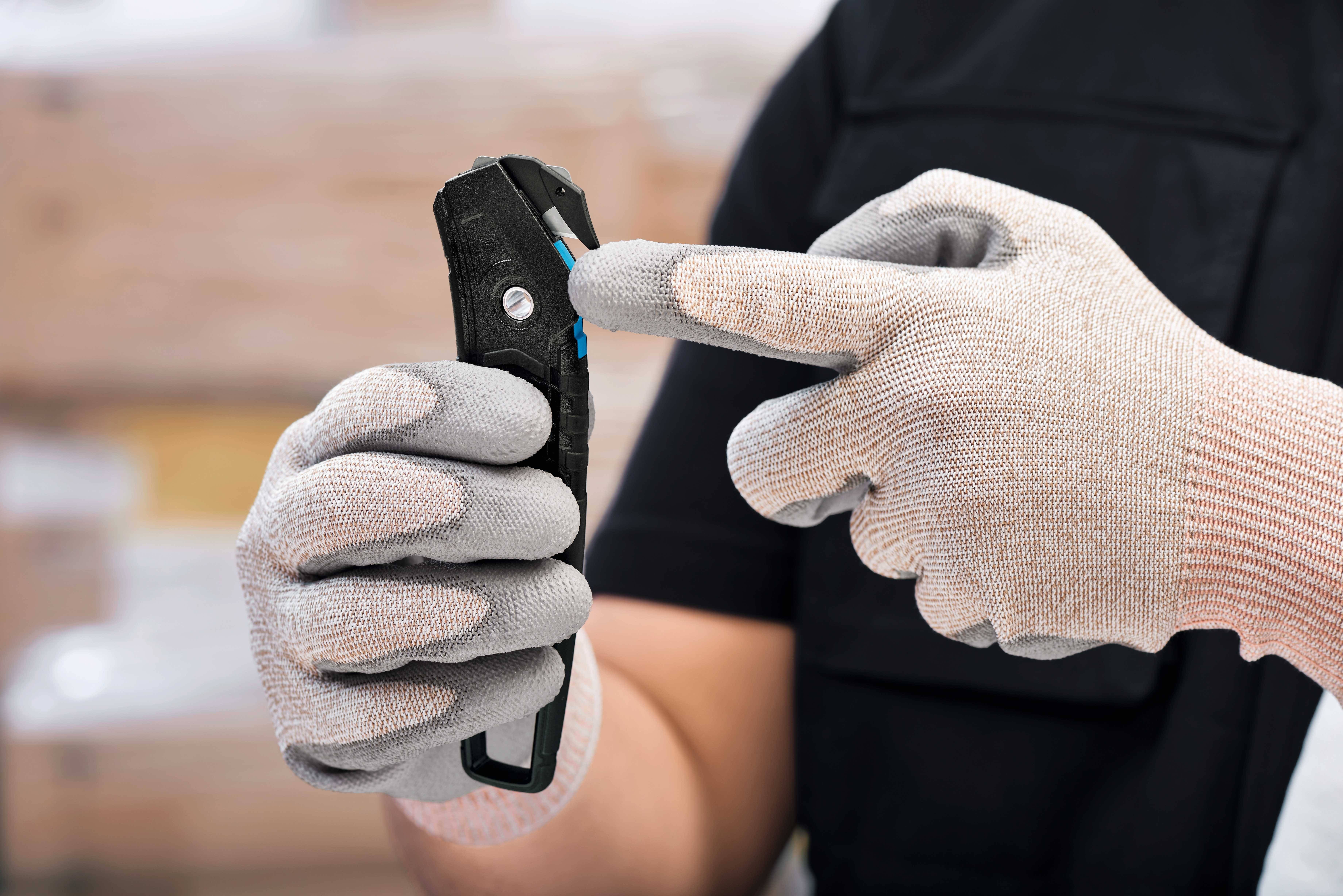 Person in Handschuhen bedient ein Sicherheitsmesser. Fokus auf Fingerposition, um Bedienung zu zeigen. Hintergrund unscharf.