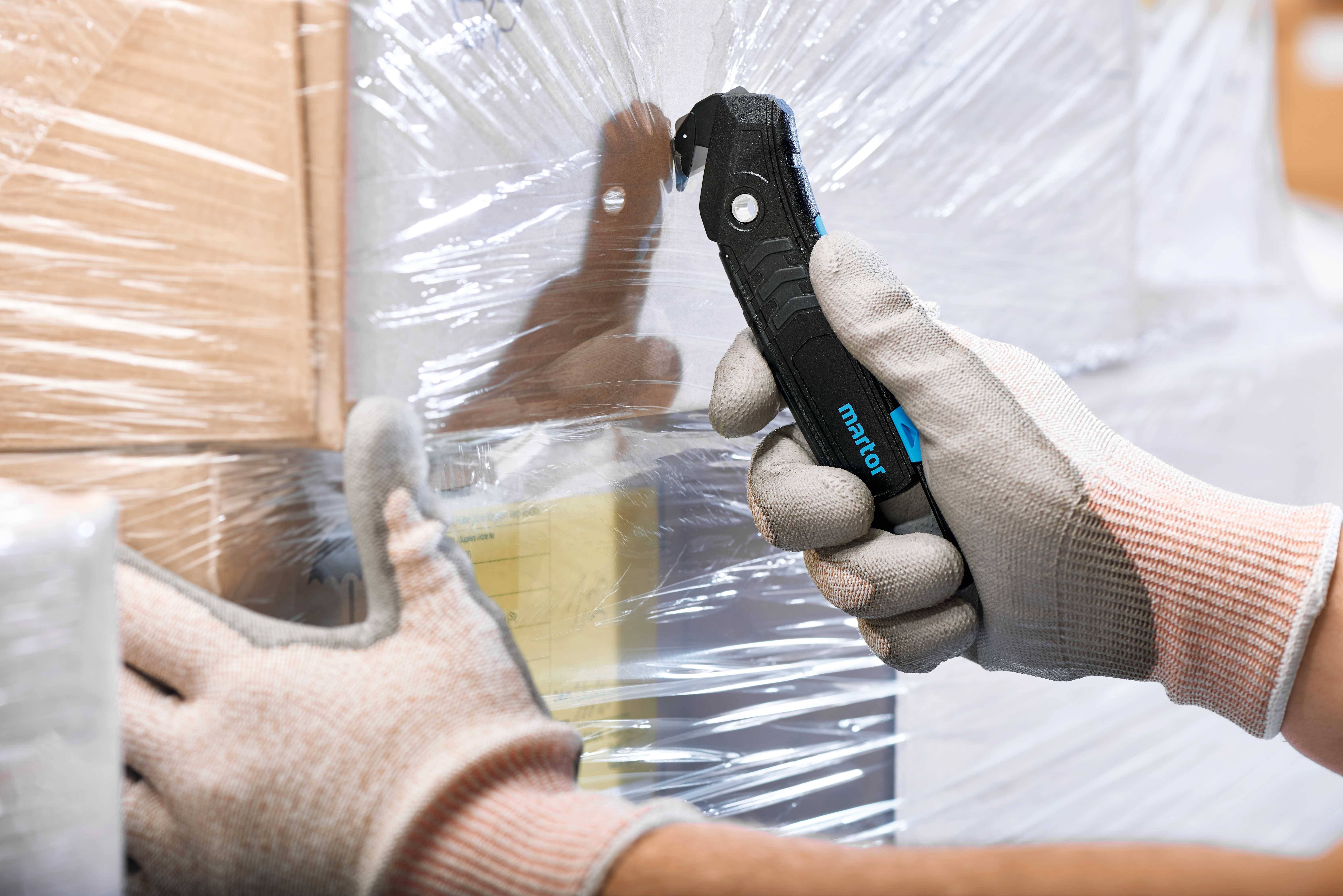 Hand zieht mit einem Sicherheitsmesser Verpackungsfolie von einem Stapel Kartons ab. Person trägt Handschuhe für zusätzlichen Schutz.