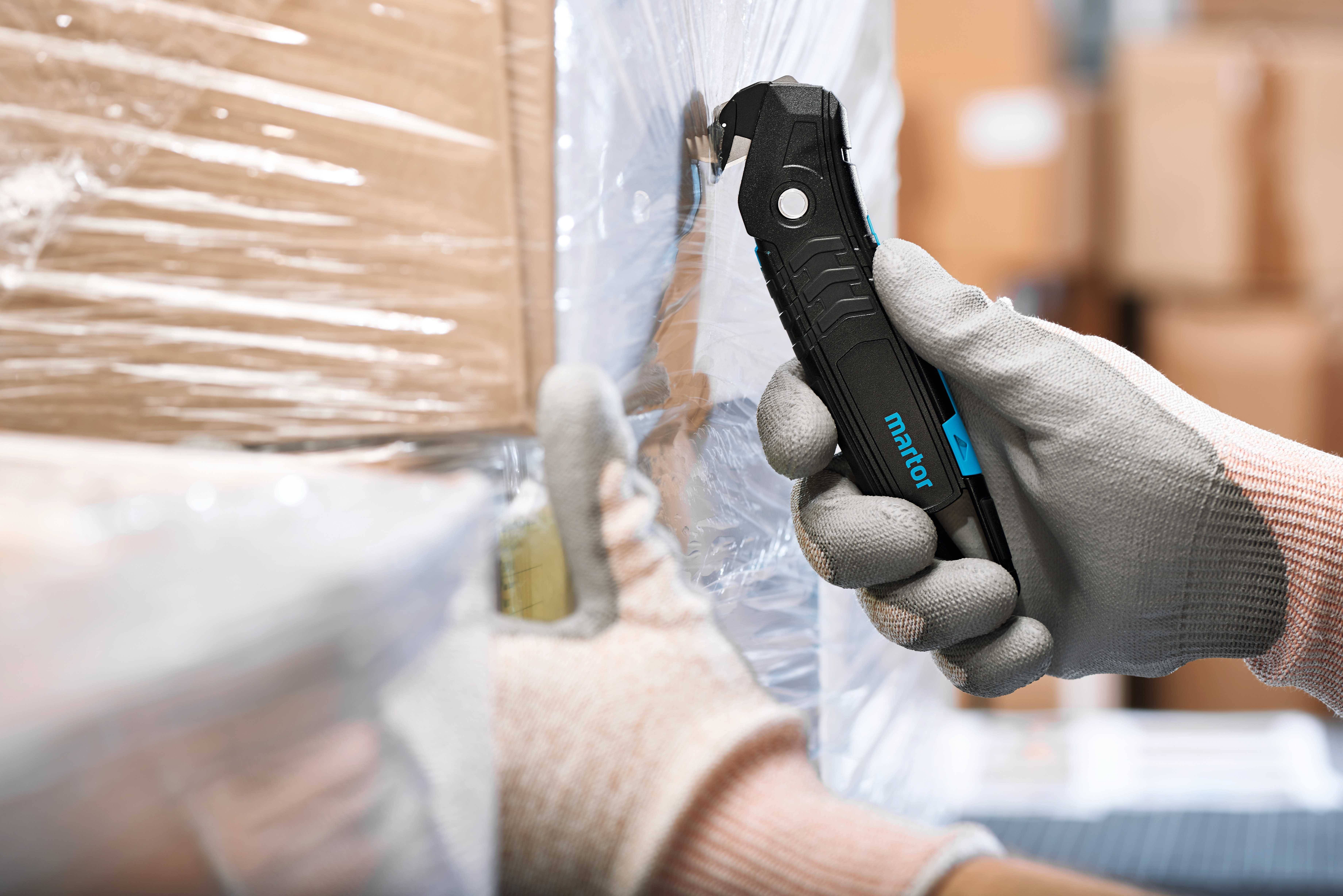 Hand in Handschuh hält ein Sicherheitsmesser; schneidet Plastikfolie von einem gestapelten Karton. Arbeitsplatz in Lagerumgebung.