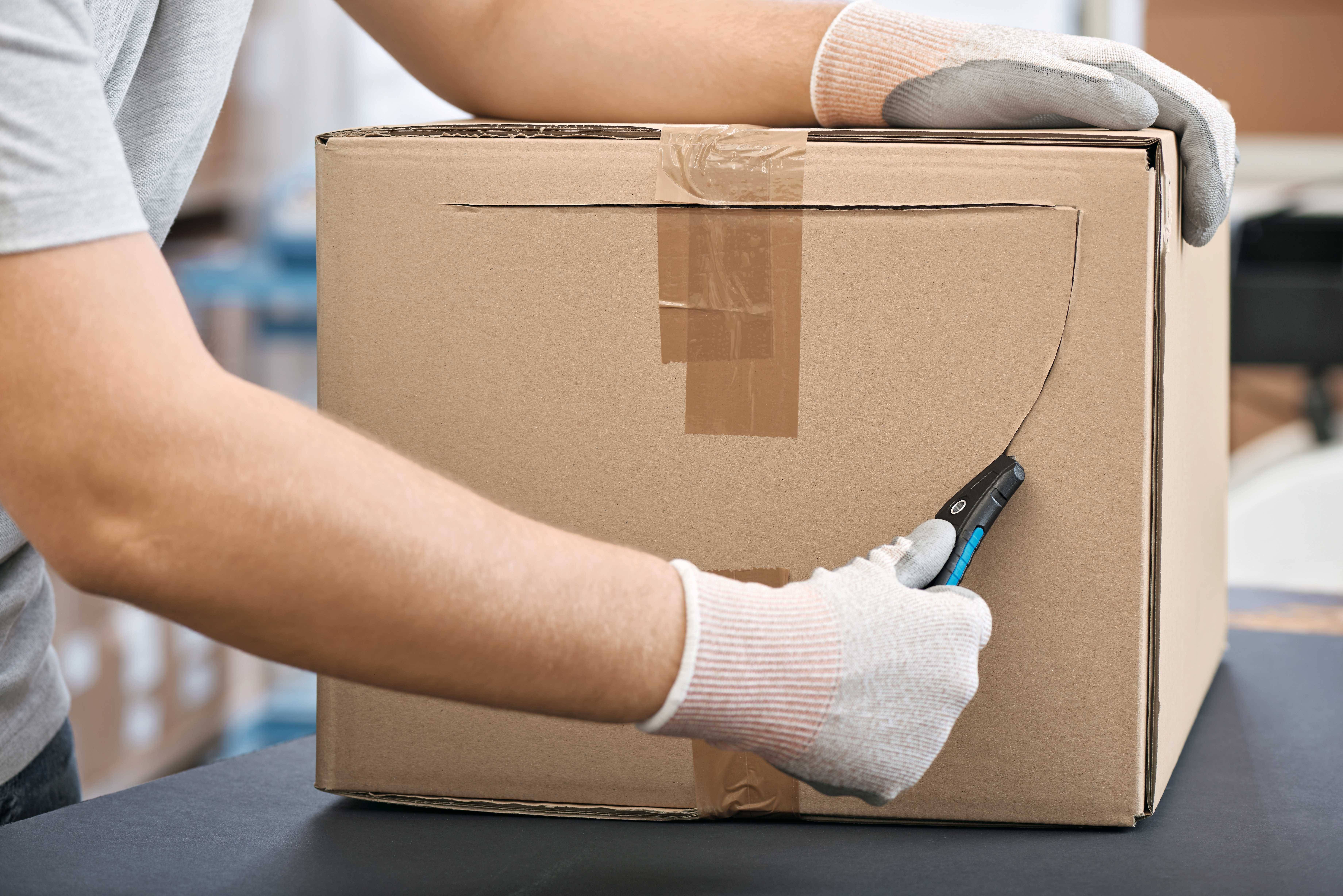 Person schneidet mit einem Cutter-Messer vorsichtig einen Karton auf. Trägt Handschuhe zum Schutz. Verpackung wird geöffnet.