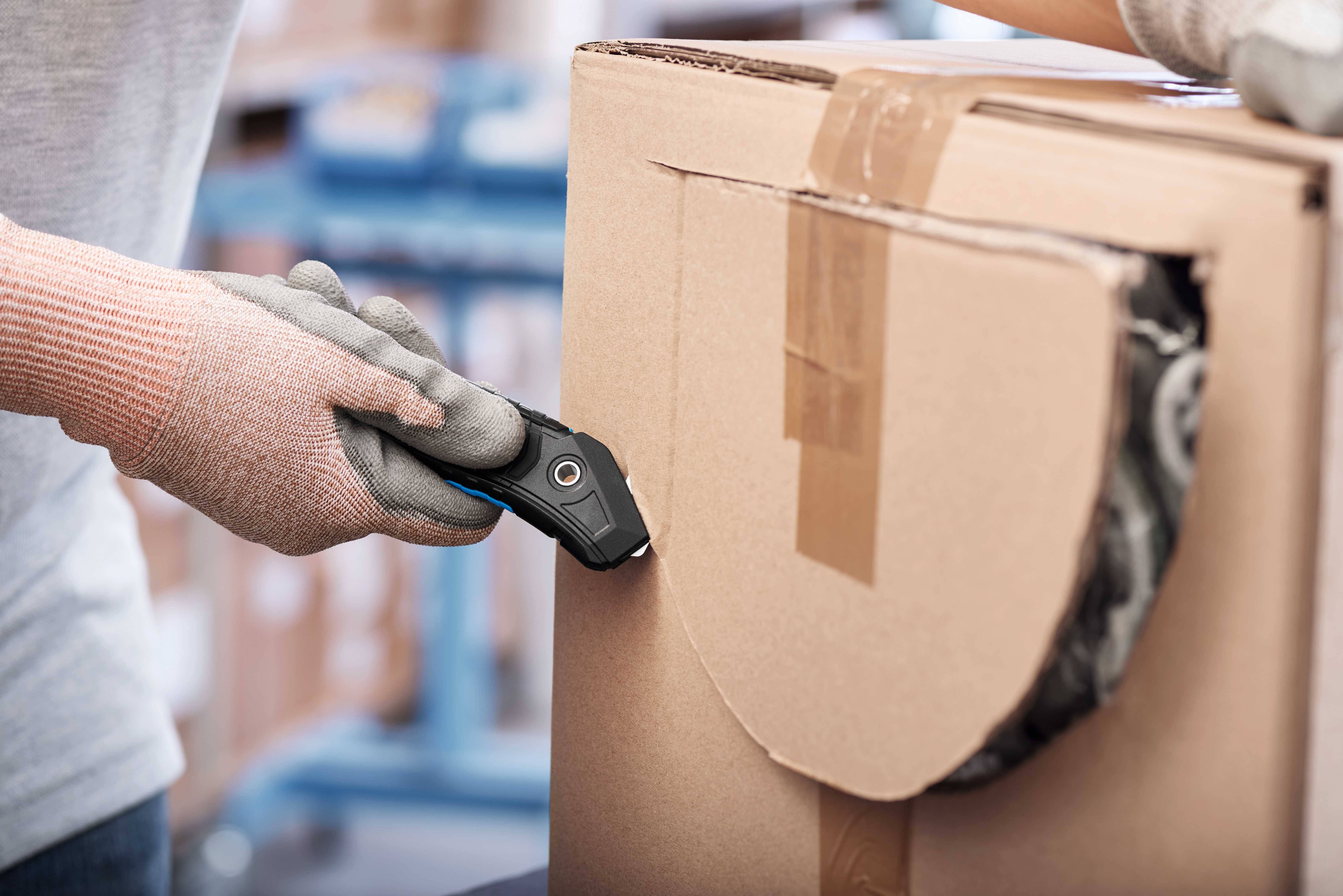 Eine Person mit Arbeitshandschuhen schneidet mit einem Sicherheitsmesser ein großes Kartonpaket auf.