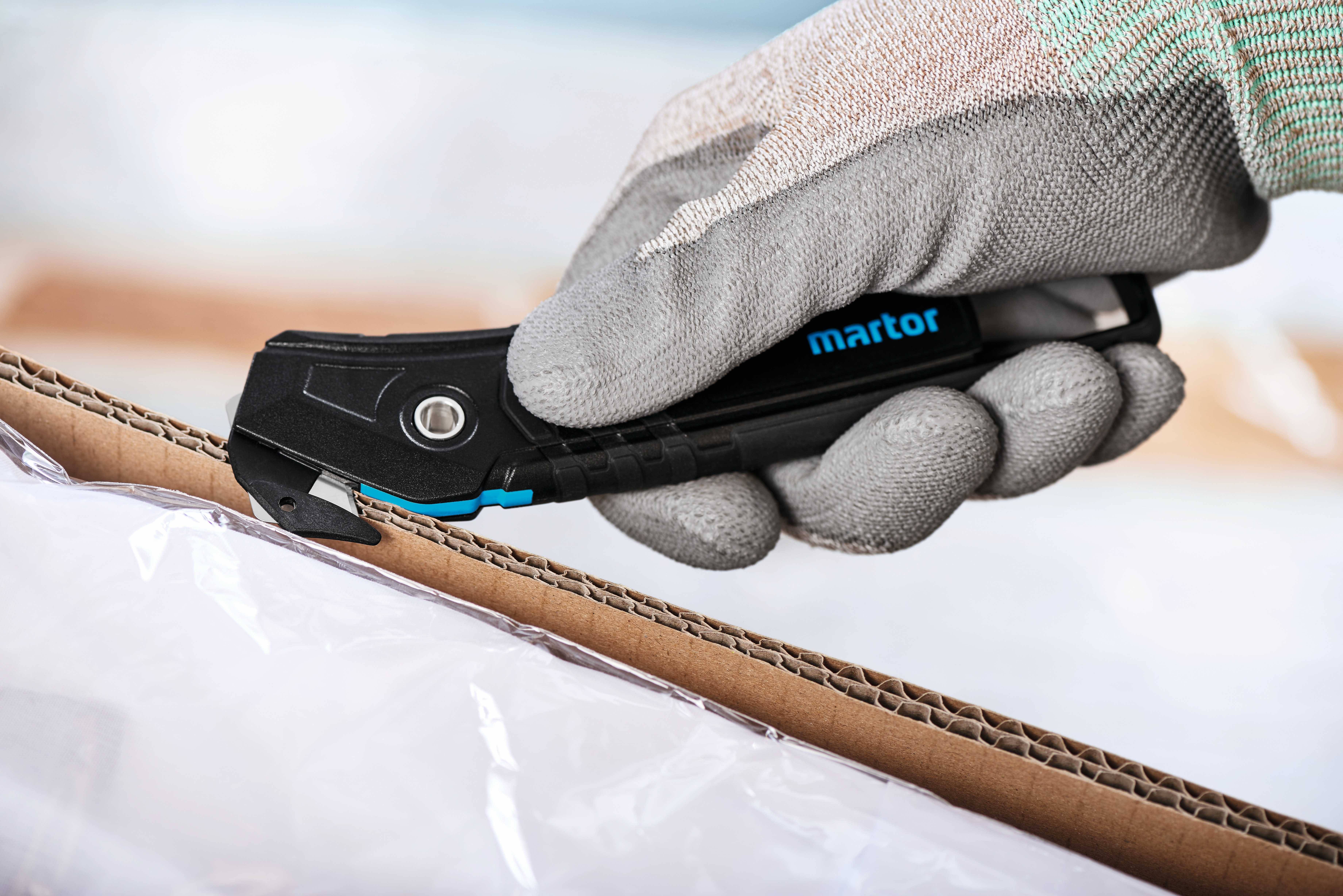 Eine behandschuhte Hand benutzt einen schwarzen Sicherheitscutter, um einen Karton zu öffnen. Der Cutter trägt den Schriftzug 'martor'.