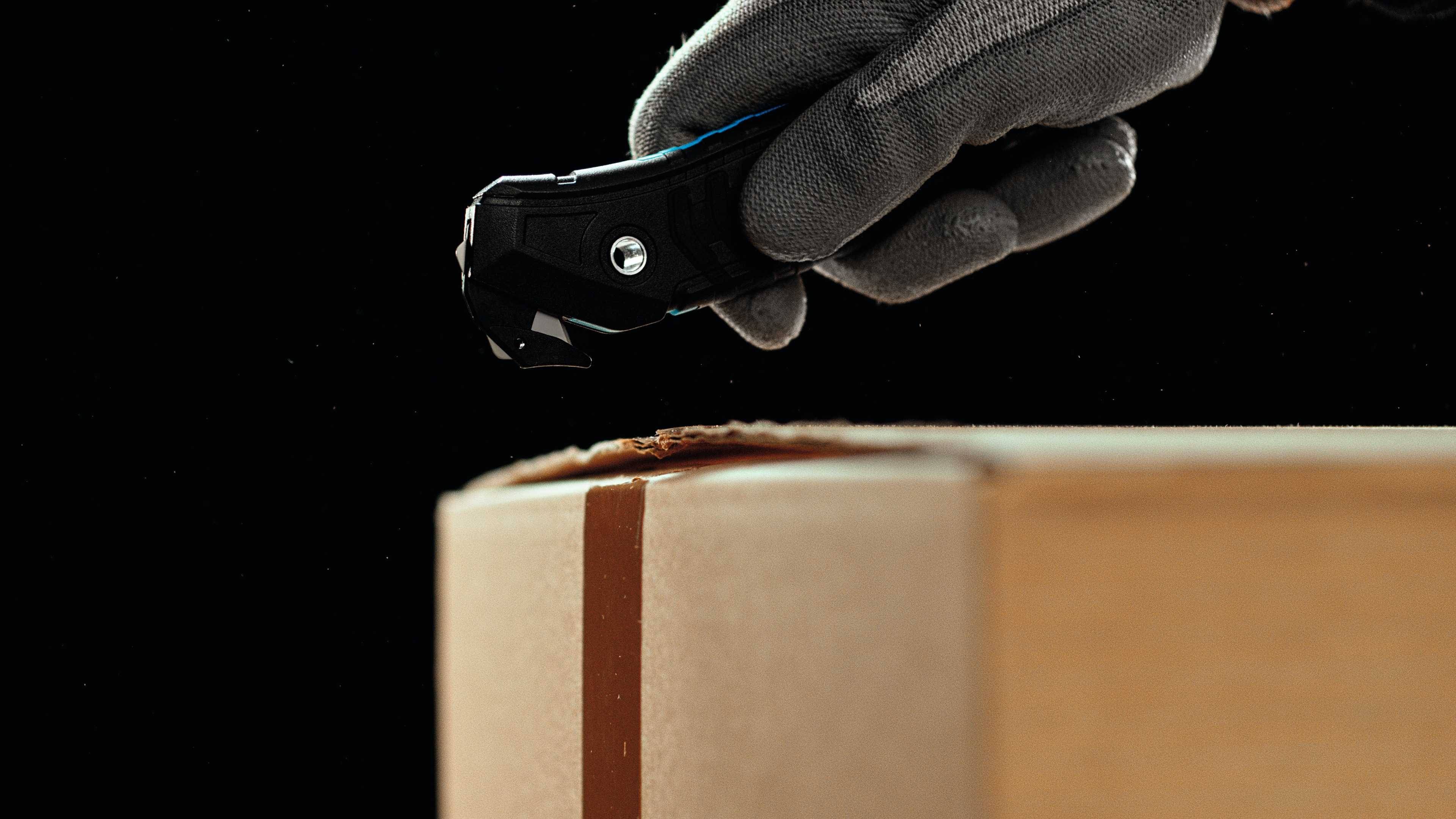 Eine Hand in einem grauen Handschuh öffnet vorsichtig einen Pappkarton mit einem Cutter-Messer vor schwarzem Hintergrund.
