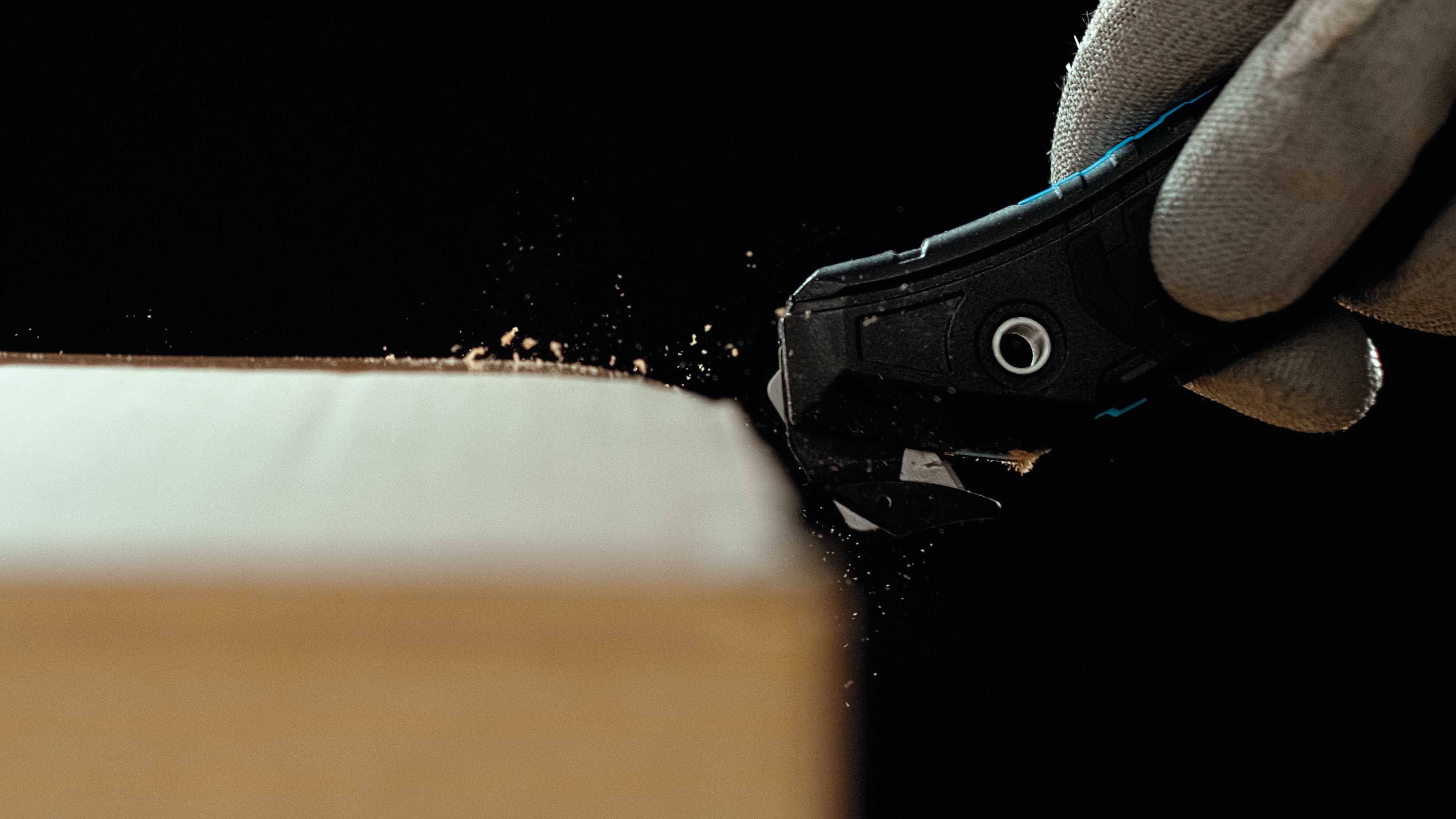 Eine behandschuhte Hand verwendet ein Teppichmesser, um Holz zu schneiden. Holzspäne fliegen, während die Klinge entlang der Kante fährt.