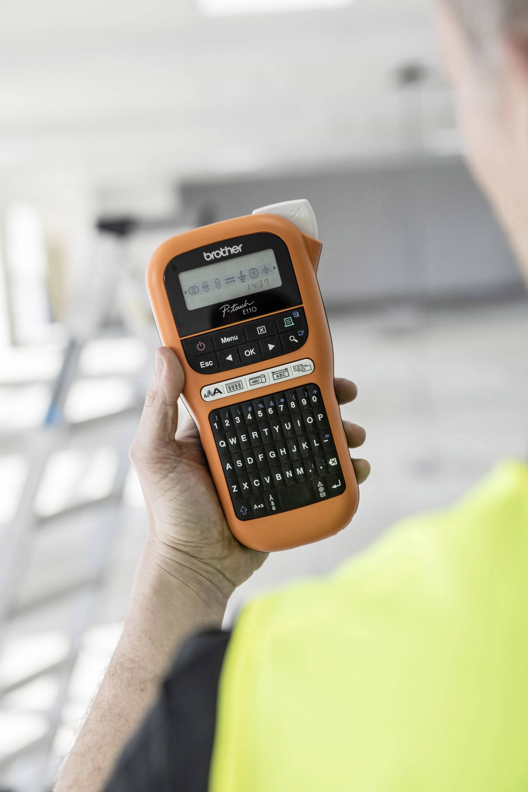 Eine Person hält ein orangefarbenes Beschriftungsgerät, das ein Display und eine Tastatur hat. Im Hintergrund ist unscharf eine Leiter sichtbar.