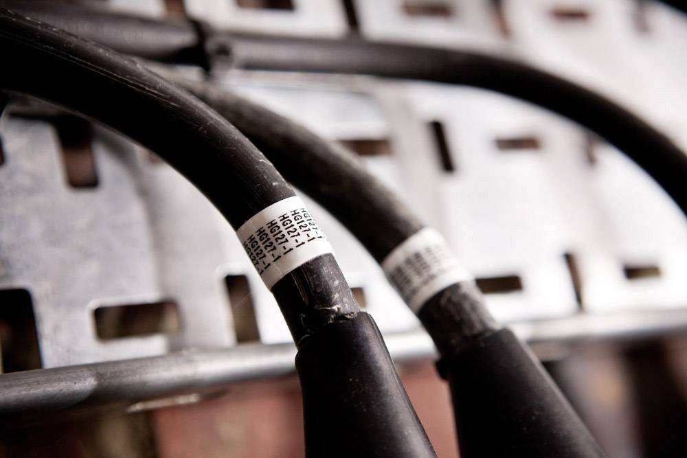 Zwei dicke schwarze Kabel mit weißen Etiketten, die auf einem Metallgitter liegen. Die Etiketten tragen unscharfe alphanumerische Codes.