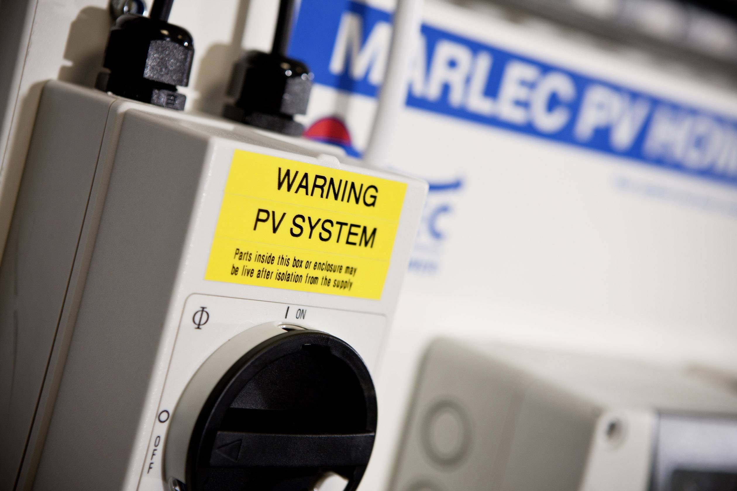Warnschild neben PV-System-Komponente mit Warnung 'WARNING PV SYSTEM'. Im Hintergrund ein unscharfer Marlec PV-Aufkleber. Hauptfokus auf Sicherheitshinweis.