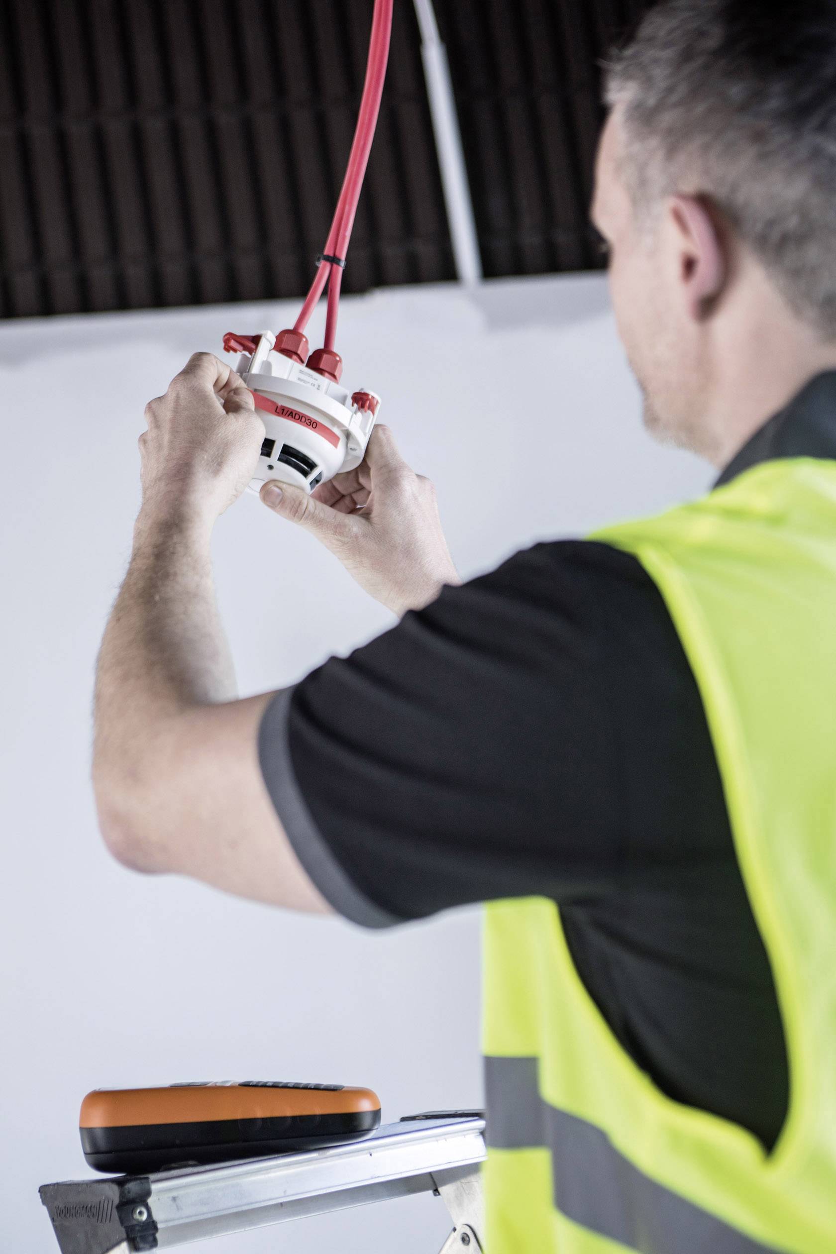 Ein Bauarbeiter in gelber Weste installiert eine Rauchmelder-Deckenmontage. Er steht auf einer Leiter und hält rote Kabel.