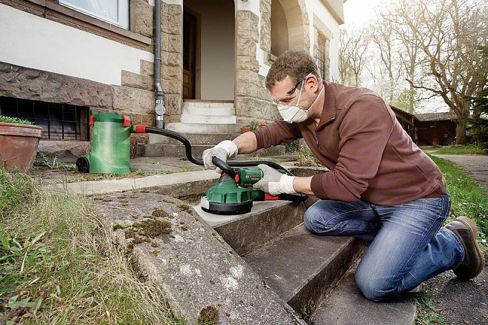 Mann mit Schutzbrille und Maske schleift Steinoberfläche auf Außentreppe. Staubsauger im Hintergrund im Garten.