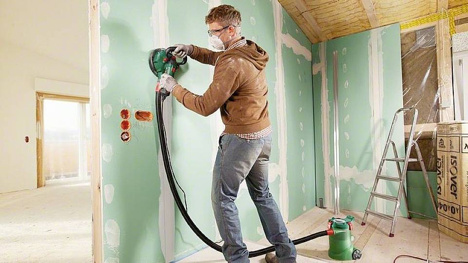 Ein Mann schleift eine grüne Trockenbauwand in einem teilweise renovierten Raum. Er trägt Schutzbrille und Staubmaske.