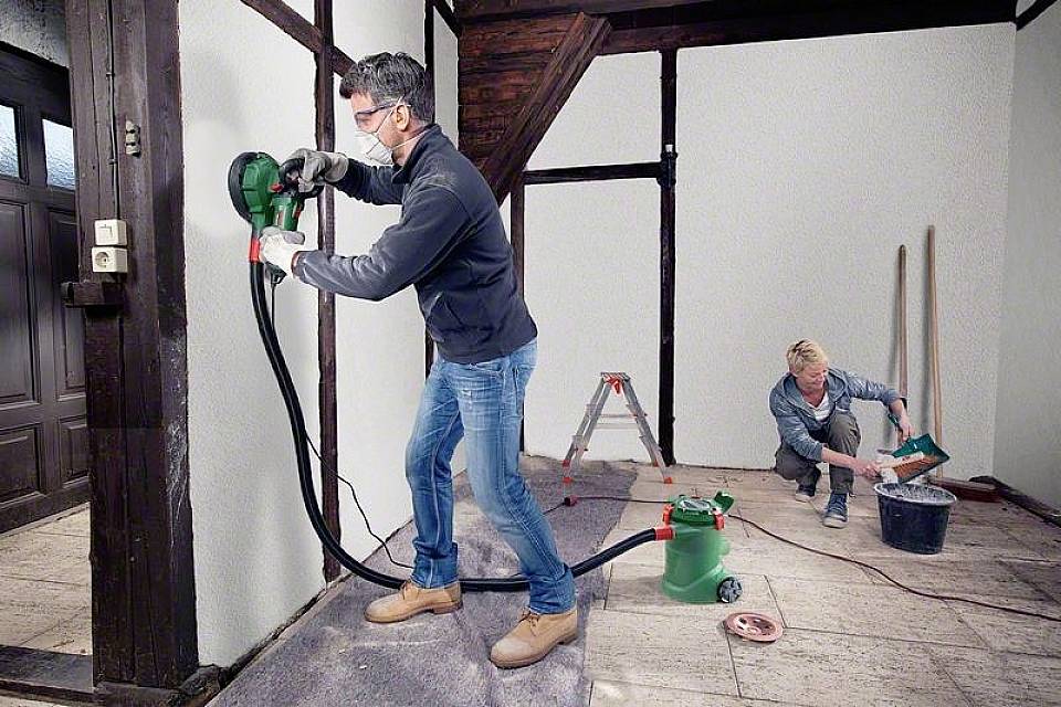 Zwei Personen renovieren in einem Raum, einer schleift die Wand mit einem Elektrowerkzeug, der andere mischt Material in einem Eimer.