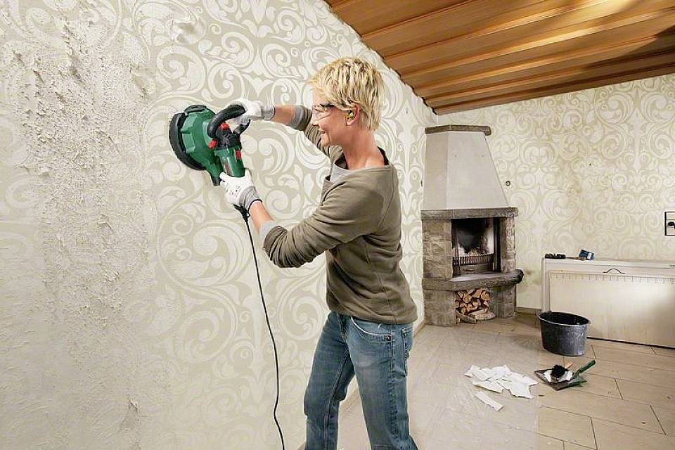 Person schleift mit einer Handschleifmaschine Tapete von der Wand in einem Raum mit Kamin. Schutzbrille und Gehörschutz werden getragen.