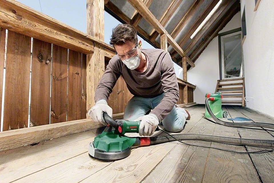 Mann schleift Holzboden auf einem Balkon. Er trägt Schutzbrille, Maske und Handschuhe. Staubsauger und Werkzeuge im Hintergrund.