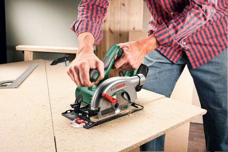 Eine Person sägt Holz mit einer grünen Handkreissäge auf einem Tisch in einer Werkstatt. Die Umgebung zeigt Sägespäne und Holzplatten.