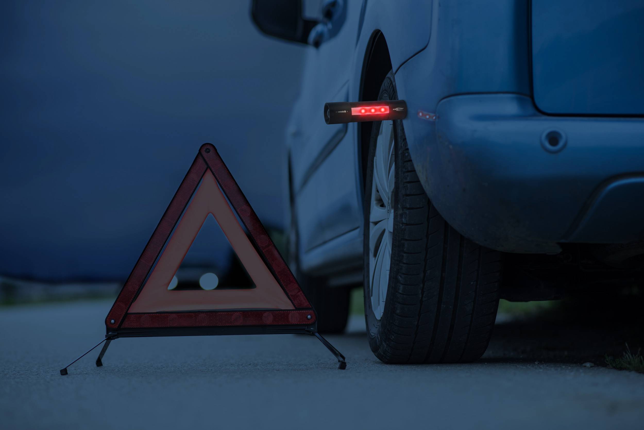 Ein blaues Auto mit einer Reifenpanne steht am Straßenrand. Ein Warndreieck mit reflektierenden Kanten steht vor dem Auto.