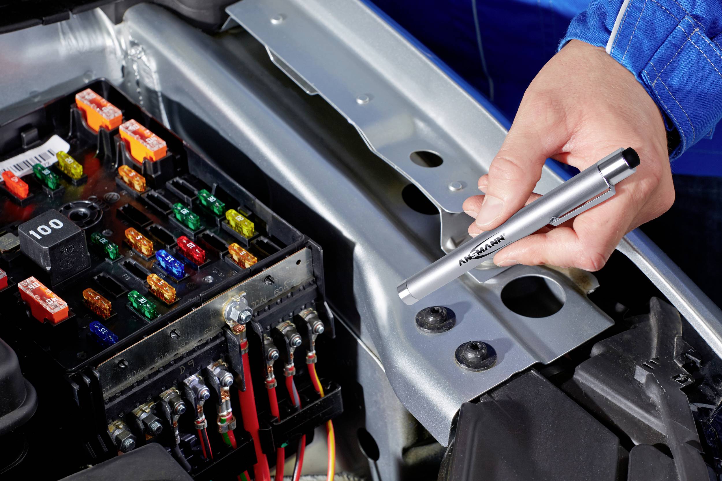 Eine Hand hält einen Spannungsprüfer über einem offenen Sicherungskasten im Motorraum eines Autos. Verschiedene farbige Sicherungen sind sichtbar.