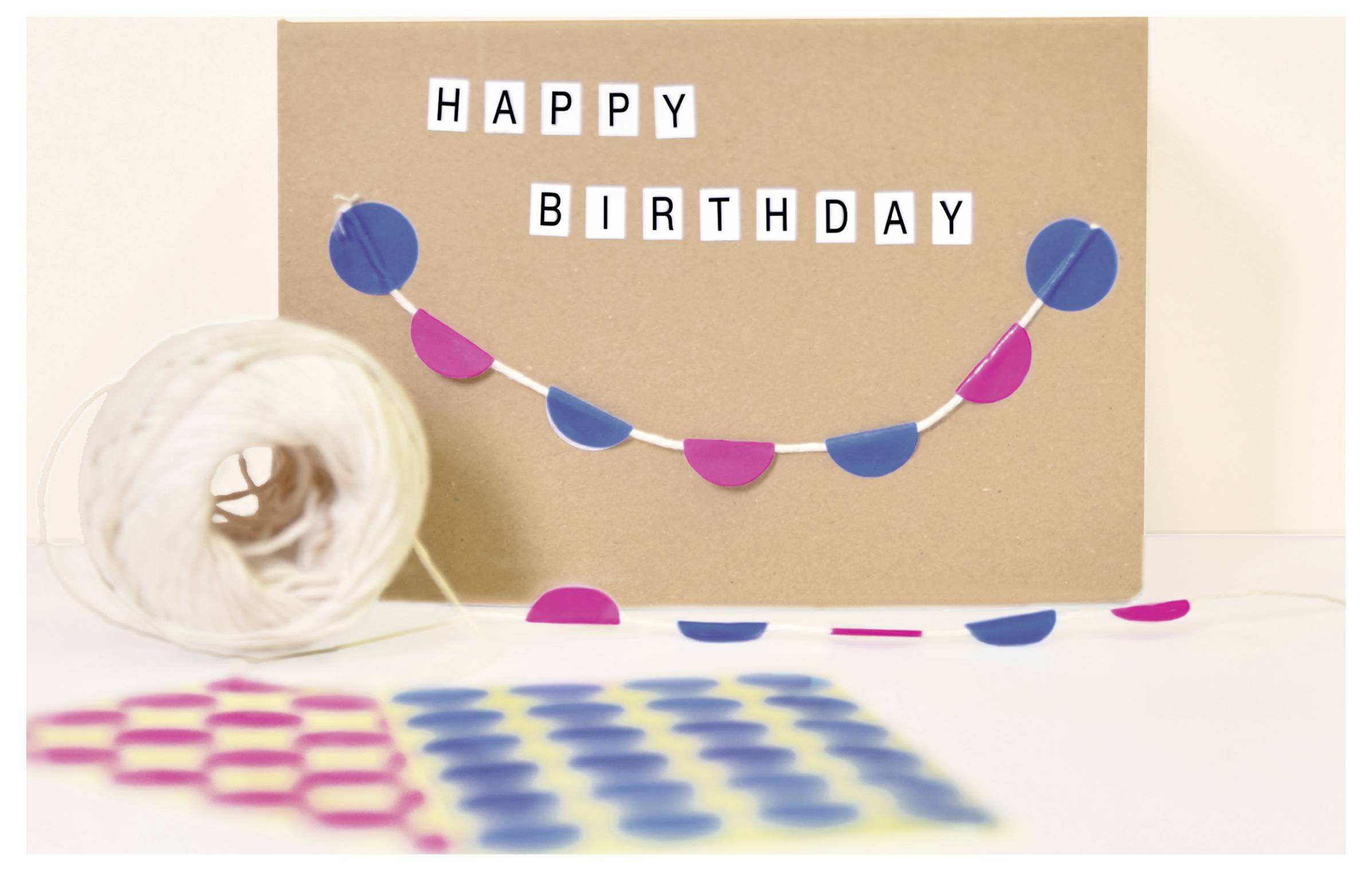 Geburtstagskarte aus Kraftpapier mit der Aufschrift 'HAPPY BIRTHDAY' und bunter Girlande; vorne liegen Papierrollen und Konfetti.