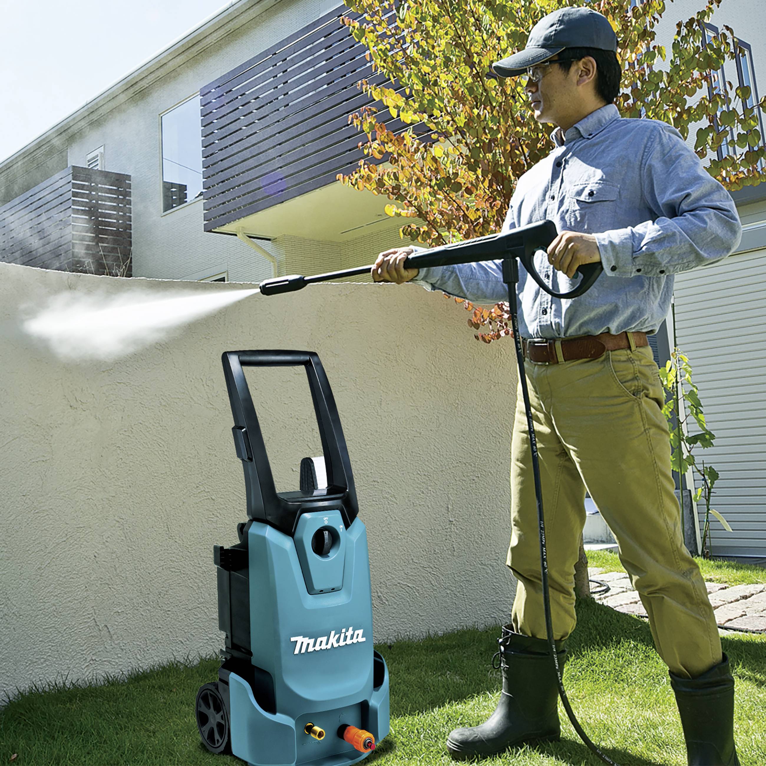 Ein Mann reinigt mit einem Hochdruckreiniger eine Außenwand. Die Sonne scheint, und im Hintergrund sind Bäume und ein modernes Gebäude zu sehen.