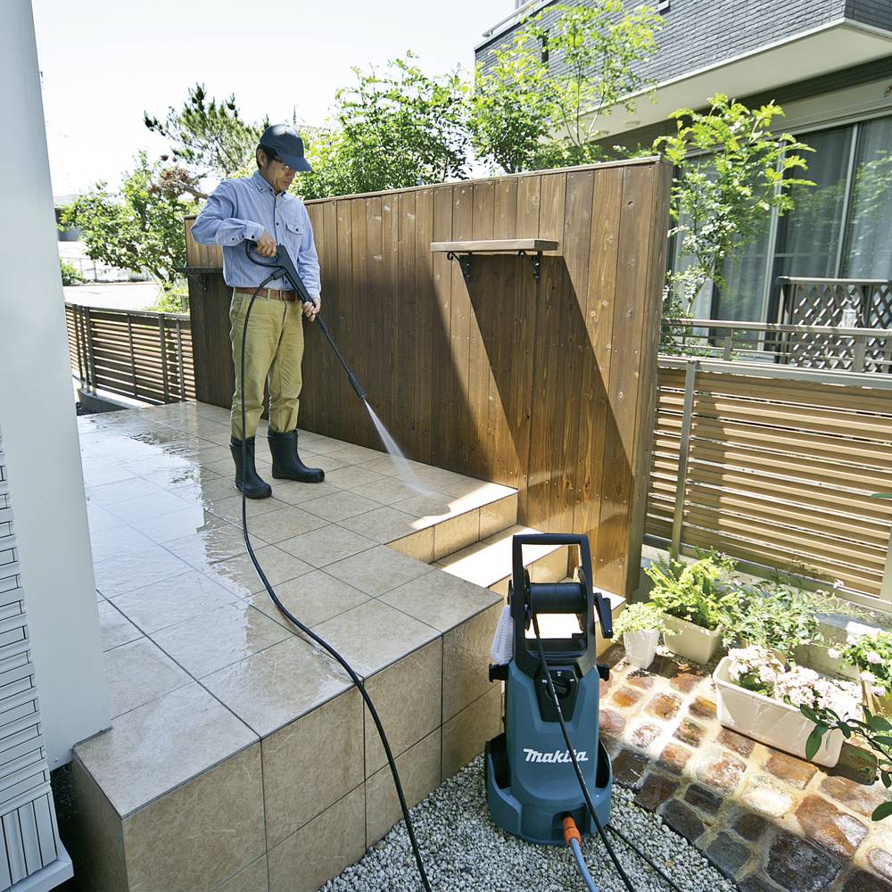 Ein Mann reinigt mit einem Hochdruckreiniger eine geflieste Terrasse. Neben ihm steht die Maschine, umgeben von Pflanzen und Zäunen.