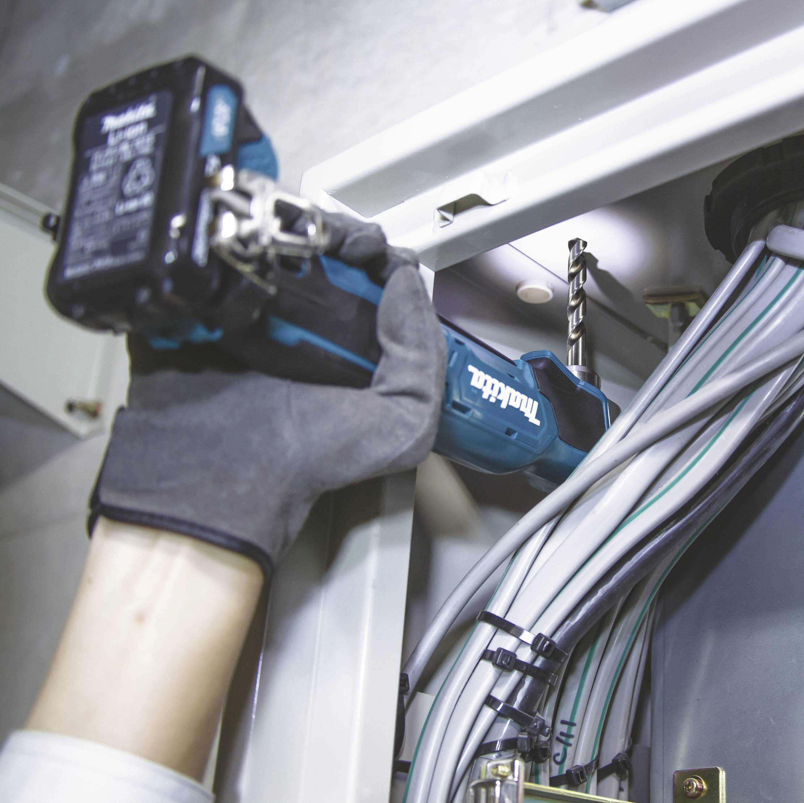 Eine Person bohrt ein Loch in eine Metallplatte. Verschiedene Kabel sind in der Nähe organisiert, was auf Elektroinstallationsarbeiten hindeutet.