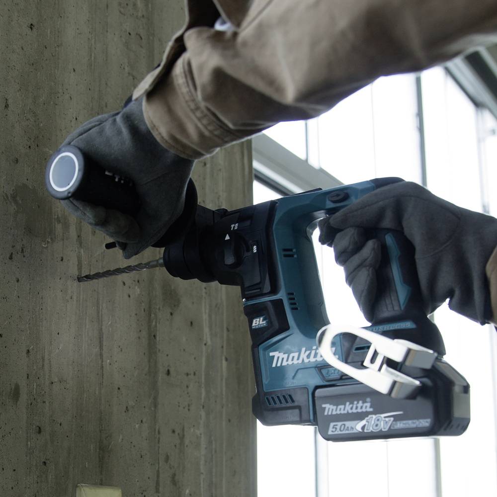 Eine Person bohrt mit einem Makita-Bohrhammer ein Loch in eine Betonwand. Im Hintergrund sind Fenster zu sehen.
